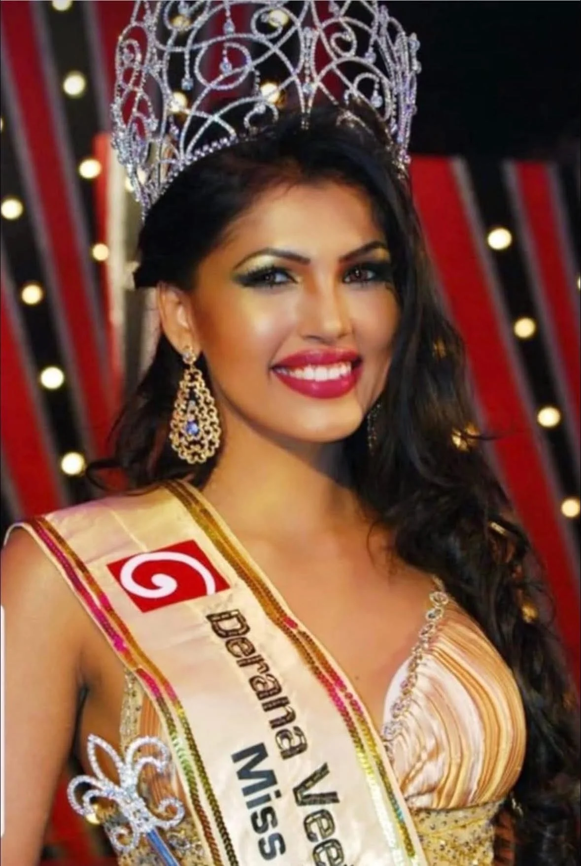 Miss Venezuela wearing a crown and sash at a beauty pageant, smiling with long dark hair, earrings, and a gold dress.