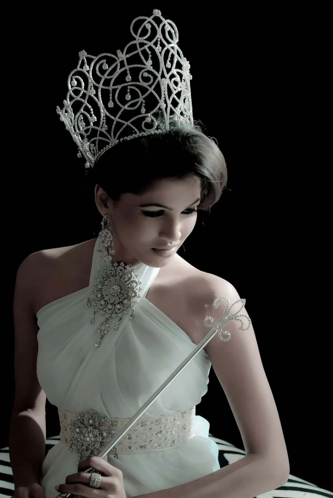 Woman wearing a white gown, ornate silver crown, and jewelry, holding a matching silver wand with a fleur-de-lis design against a black background.