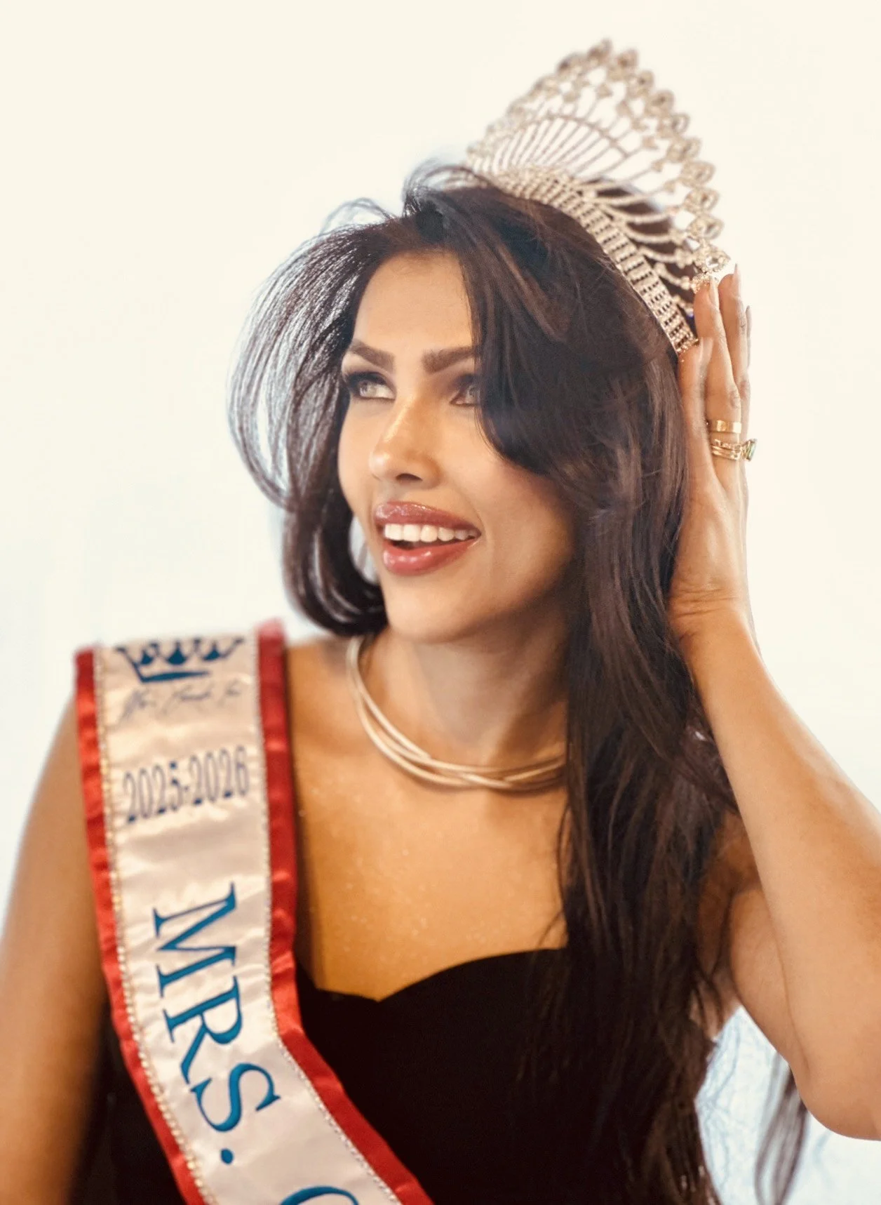A woman wearing a Miss Universe sash and crown, smiling and touching her head, dressed in black and wearing jewelry.