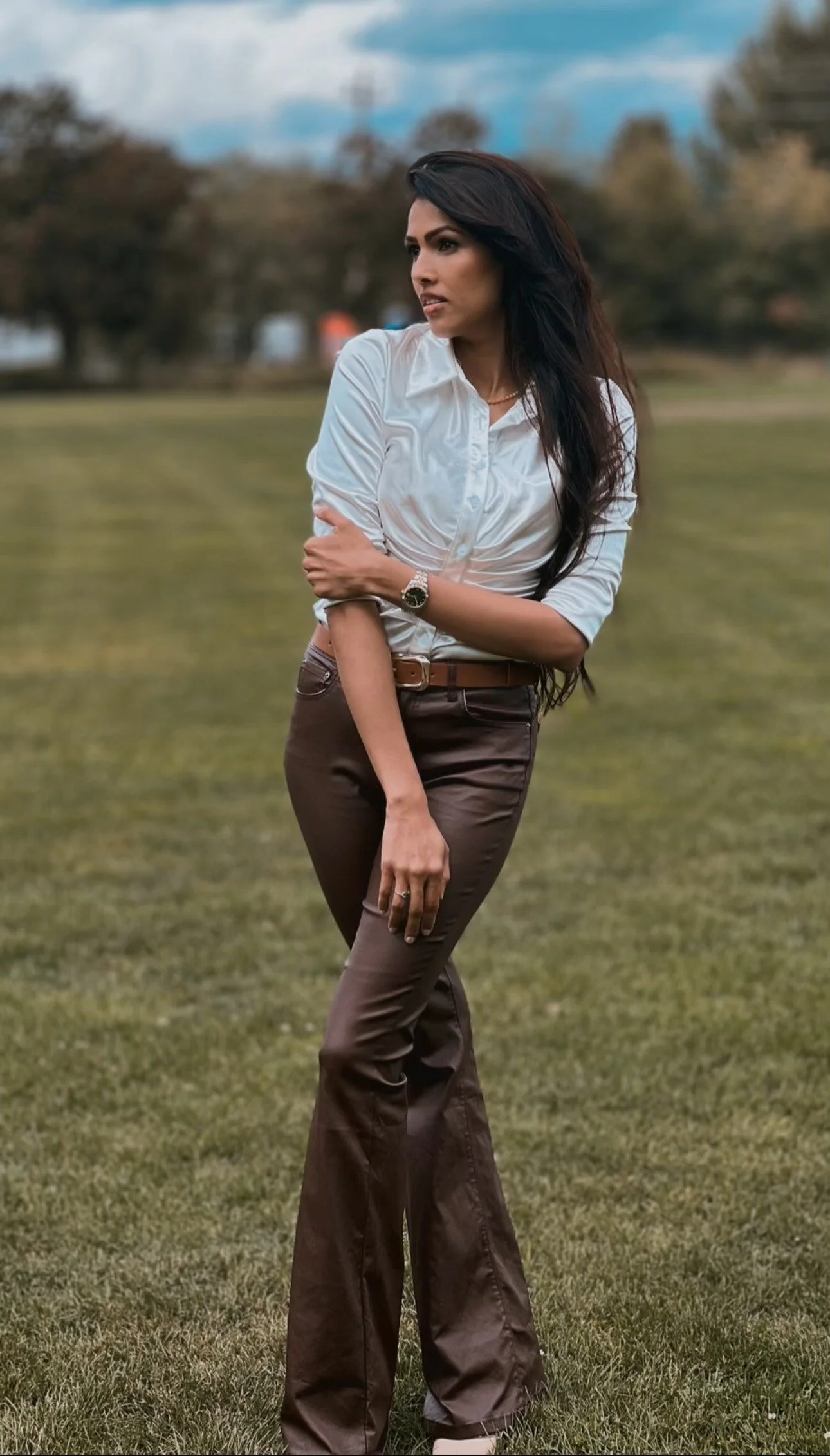 A woman standing outdoors on grass, wearing a white button-up shirt and brown pants, with dark, styled hair, looking off to the side.