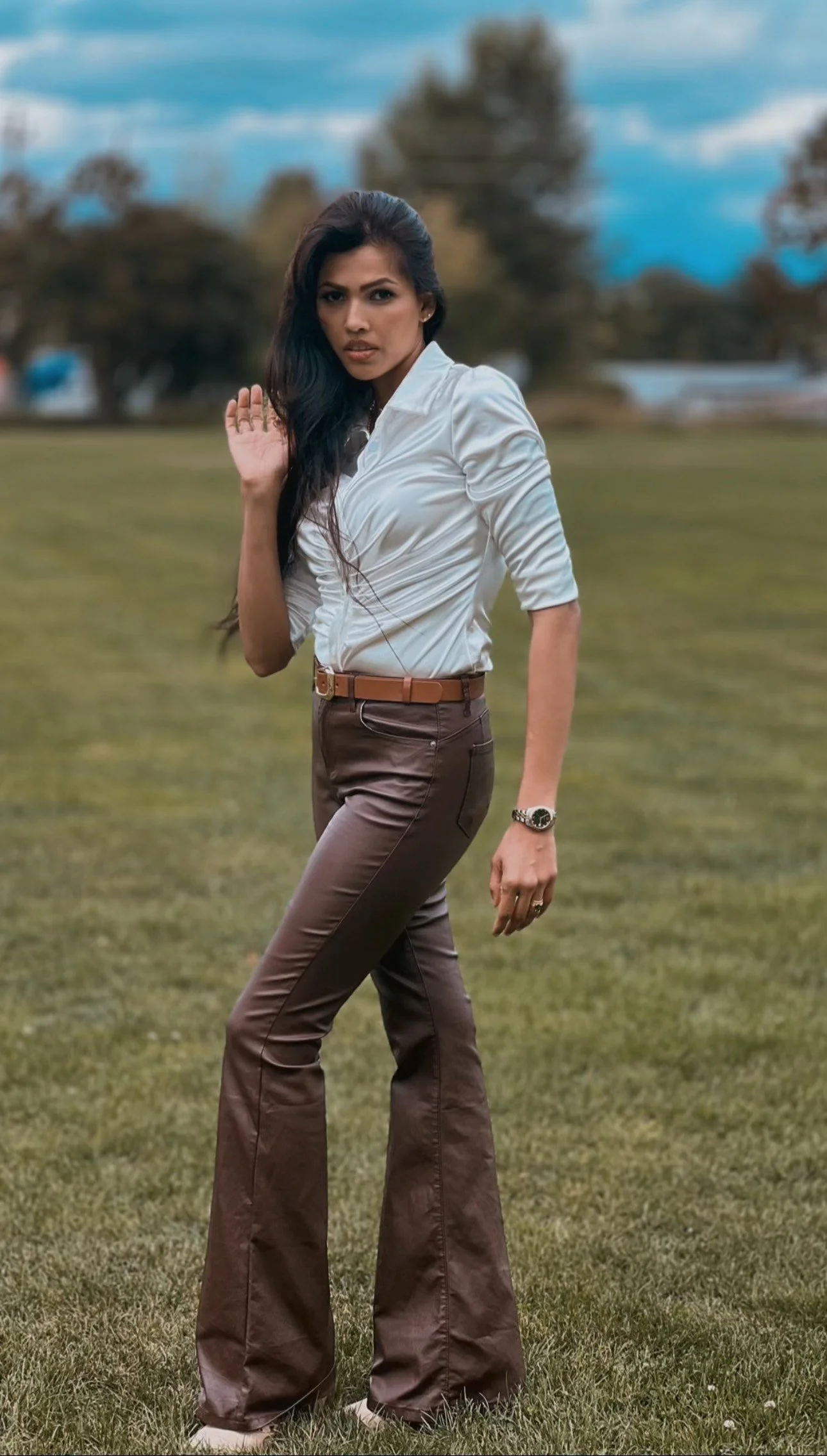 A woman with long dark hair wearing a white blouse and brown flared pants standing on a grassy field, with trees and cloudy sky in the background.