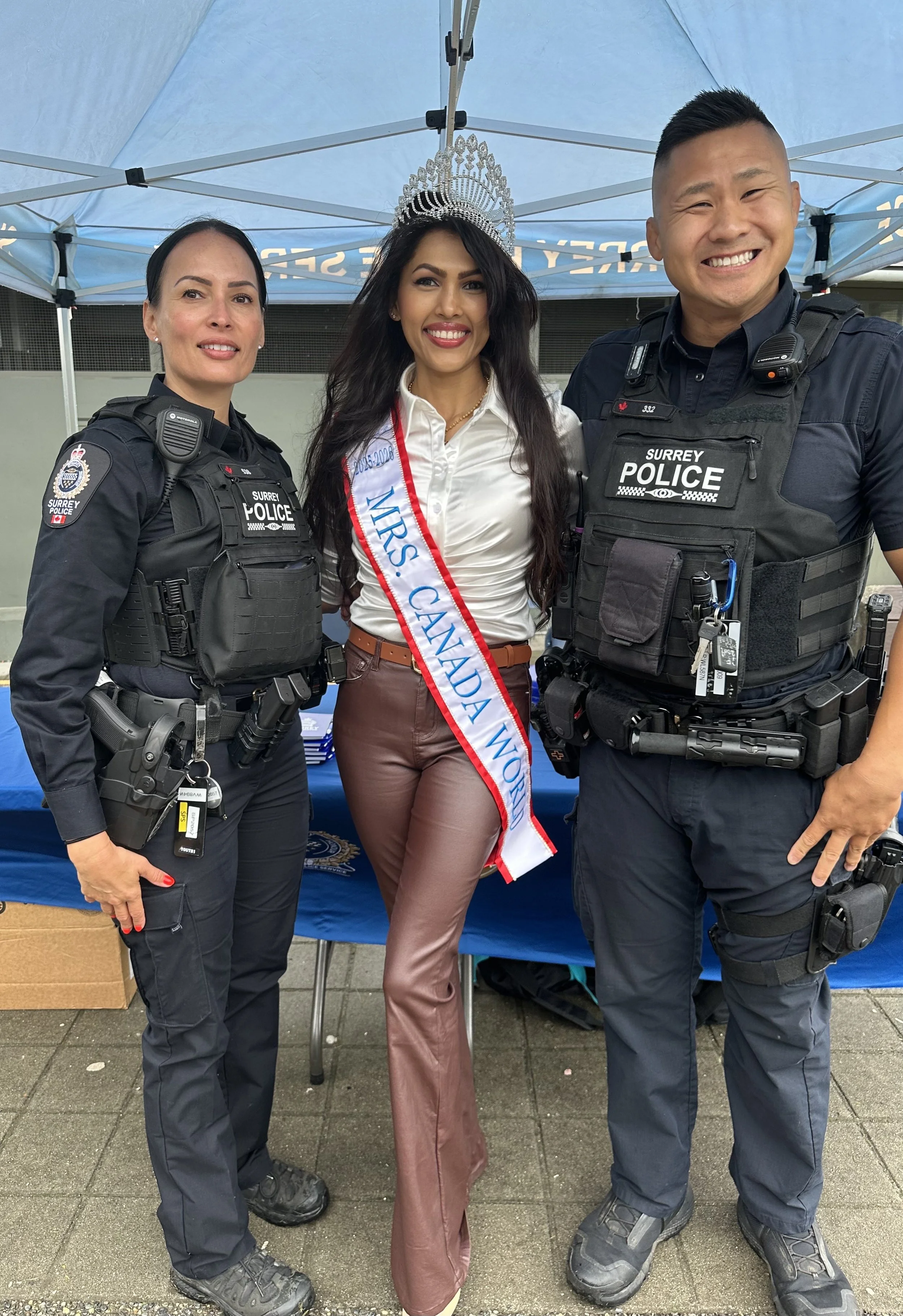 A woman wearing a tiara and sash standing between two police officers under a blue tent.