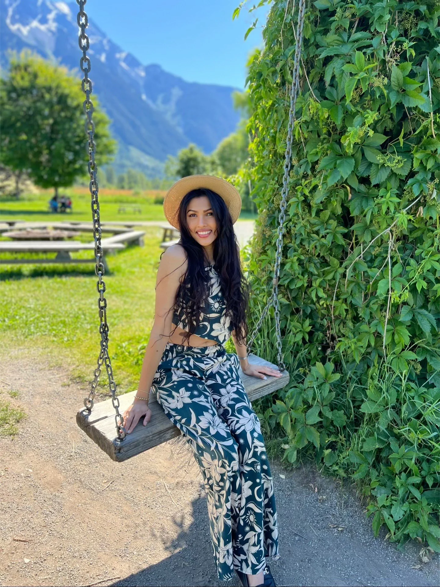 A woman in a floral summer outfit and straw hat sitting on a wooden swing outdoors, with green foliage and mountains in the background.