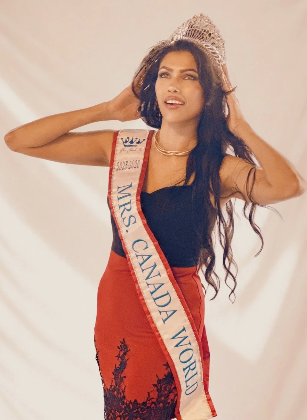A woman wearing a crown and sash that reads 'Mrs. Canada World 2025-2026' stands against a plain backdrop, wearing a black top, red skirt, and layered necklaces, with her hands adjusting her hat.