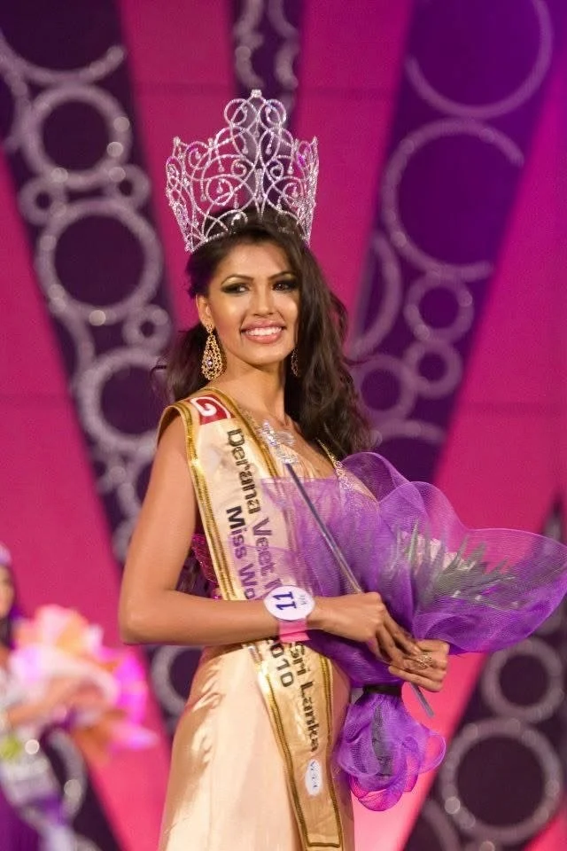 Beauty queen wearing a crown and sash, holding a bouquet of flowers, on stage during a beauty pageant.