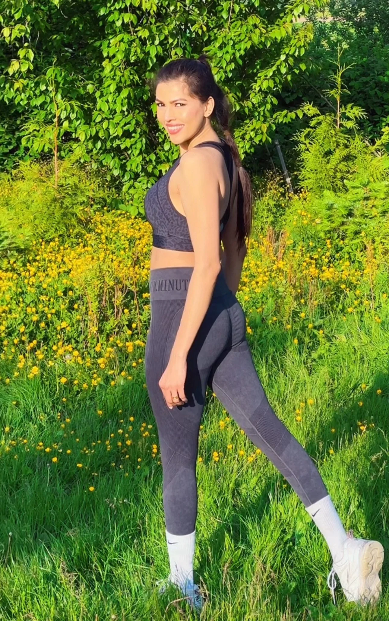 A woman in athletic wear standing outdoors in a grassy field with yellow flowers, smiling and squinting in the sunlight.