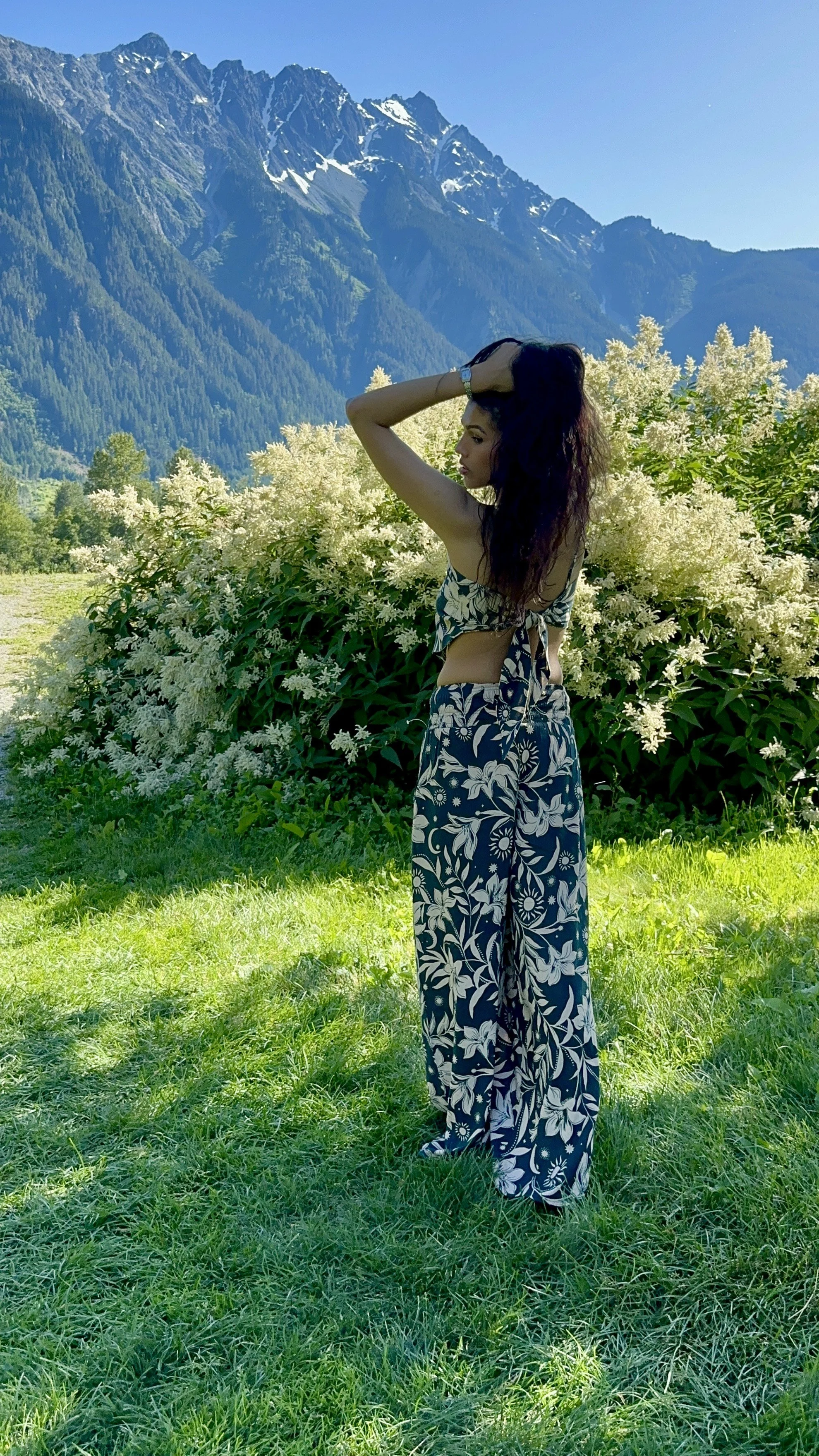 A woman with long dark curly hair standing in a green field surrounded by blooming white flowers, with a backdrop of tall, rugged mountains with patches of snow under a clear blue sky.
