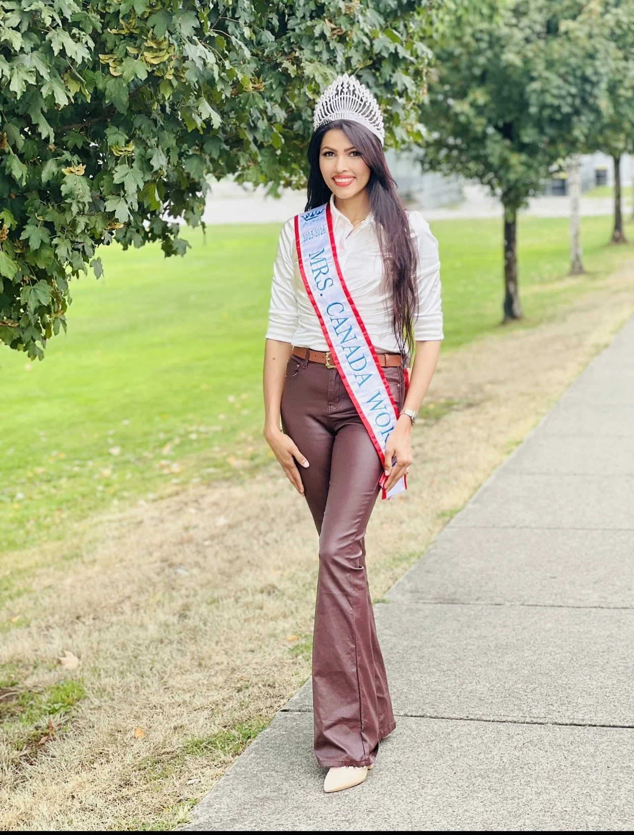 Woman wearing a crown and sash with 'Mrs. Canada World' standing outdoors on a sidewalk beside grass and trees, smiling.