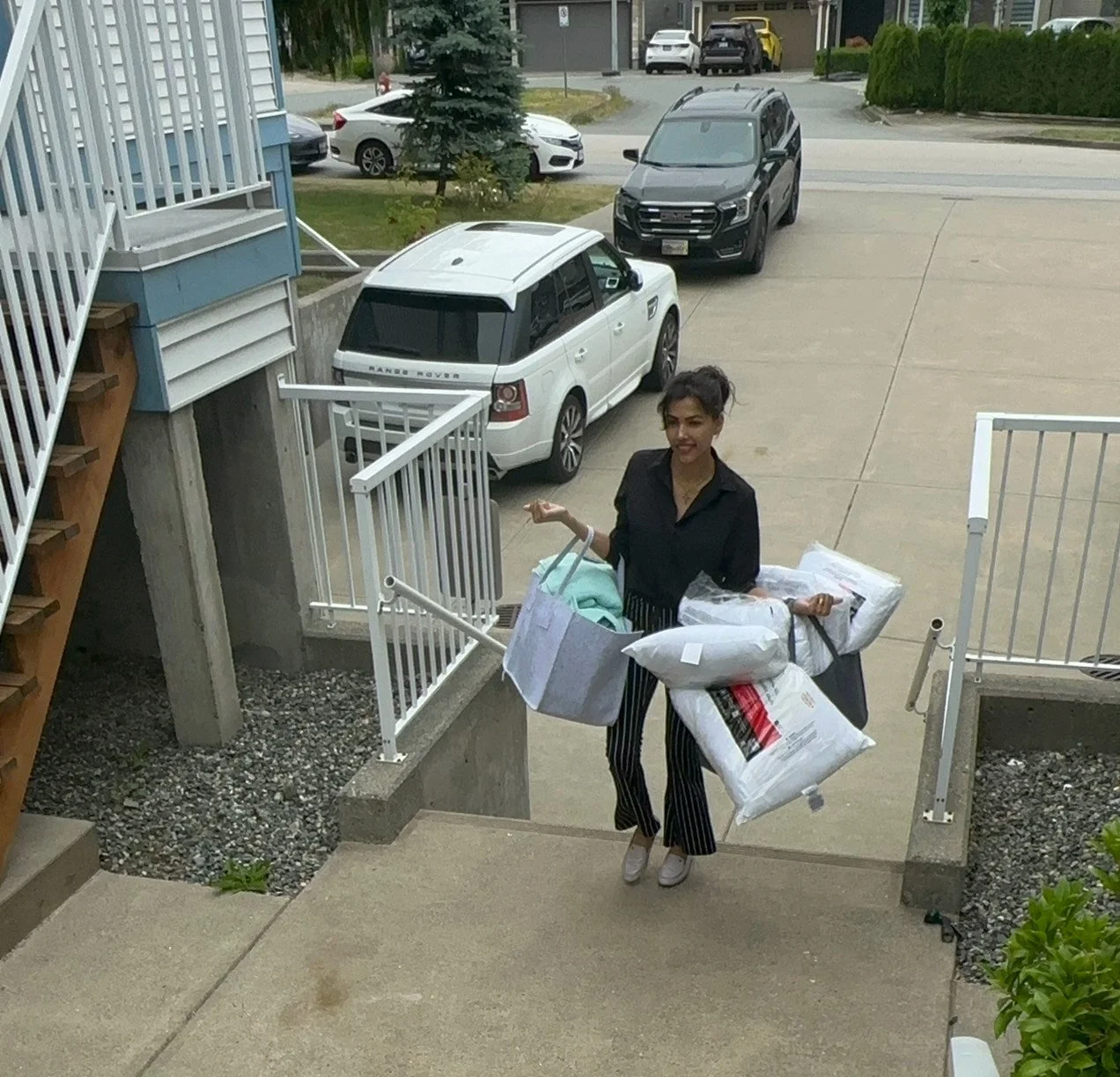 Woman carrying multiple bags and pillows on her arms, walking up stairs from an apartment building with parked cars and street in background.