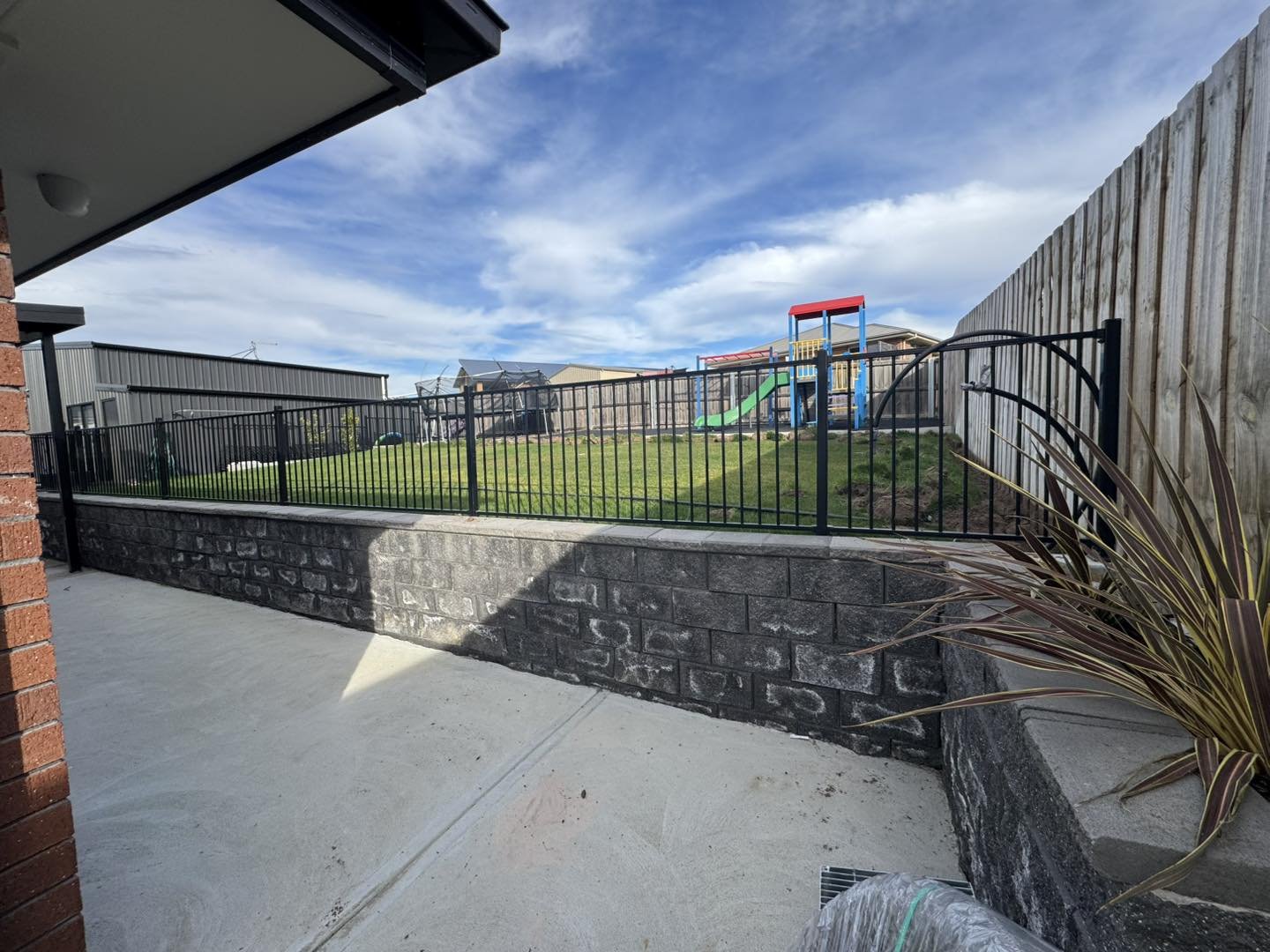 View of a backyard with a black metal fence, green grass, and a playset with slide and climbing structure under a partly cloudy sky.