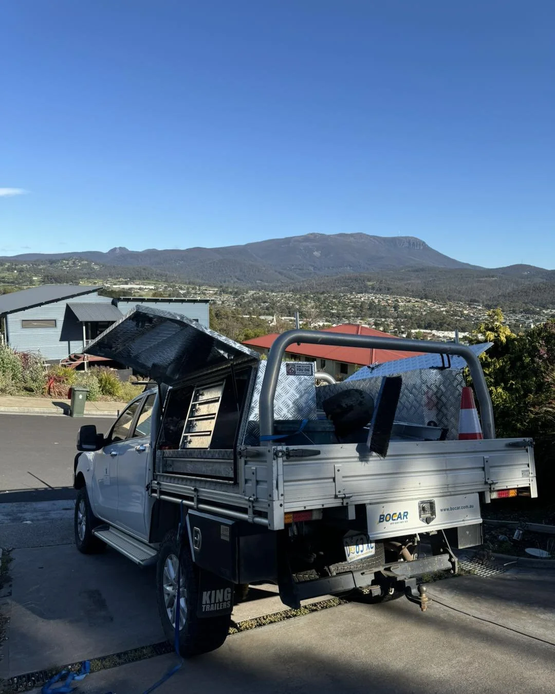 A white utility truck with an open tailgate parked on a driveway, with tools and cones visible in the truck bed, overlooking a mountainous landscape under a clear blue sky.