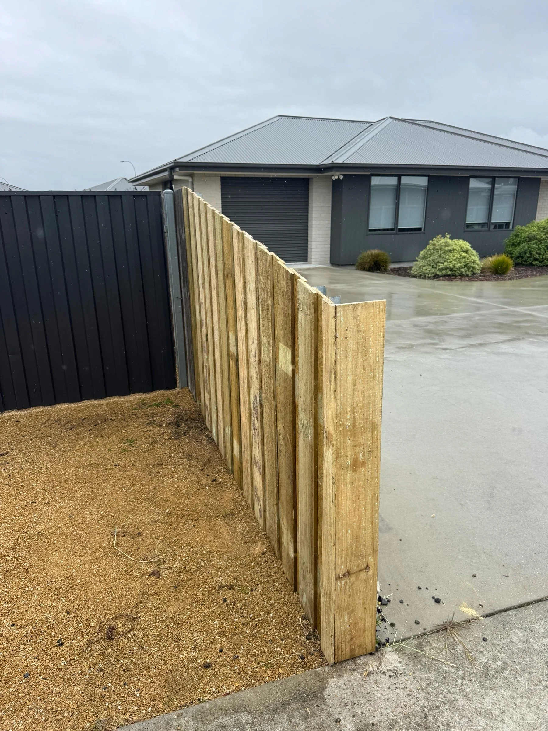 A newly built wooden fence section in front of a modern residential house with a gray metal roof and dark window frames, on a bright, overcast day.