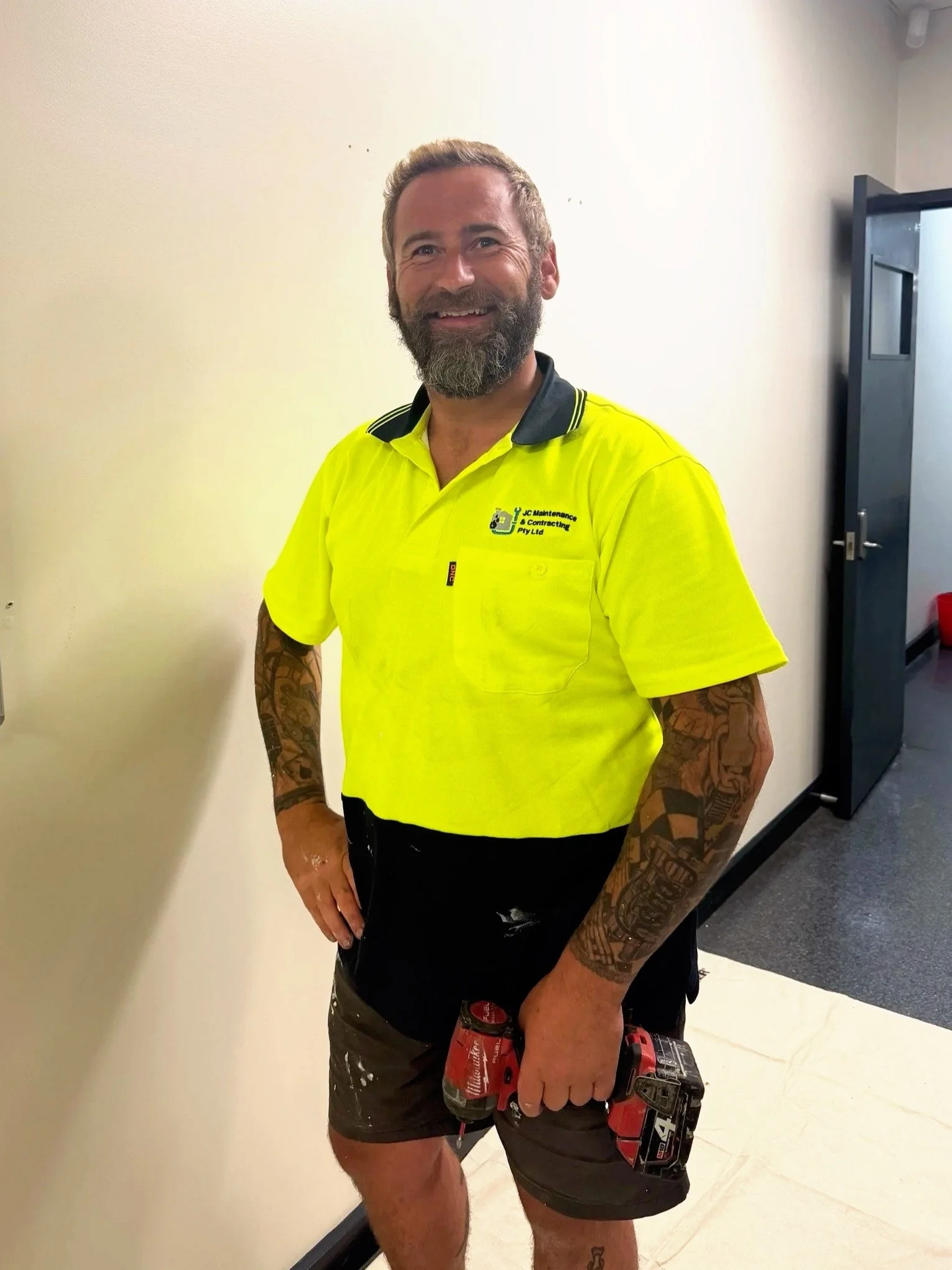 A smiling man with a beard and tattoos wearing a yellow work shirt holding a power drill, standing indoors near a white wall and an open door.