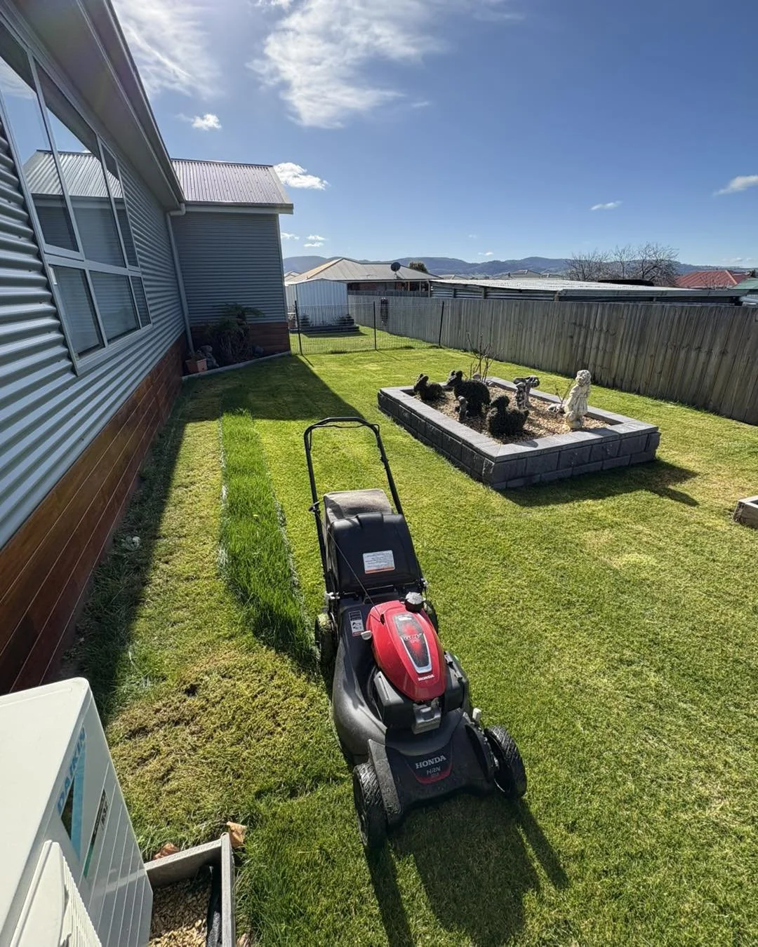 A backyard with a lawnmower, garden bed with black figurines, a house with siding, wooden fence, and distant mountains under a blue sky