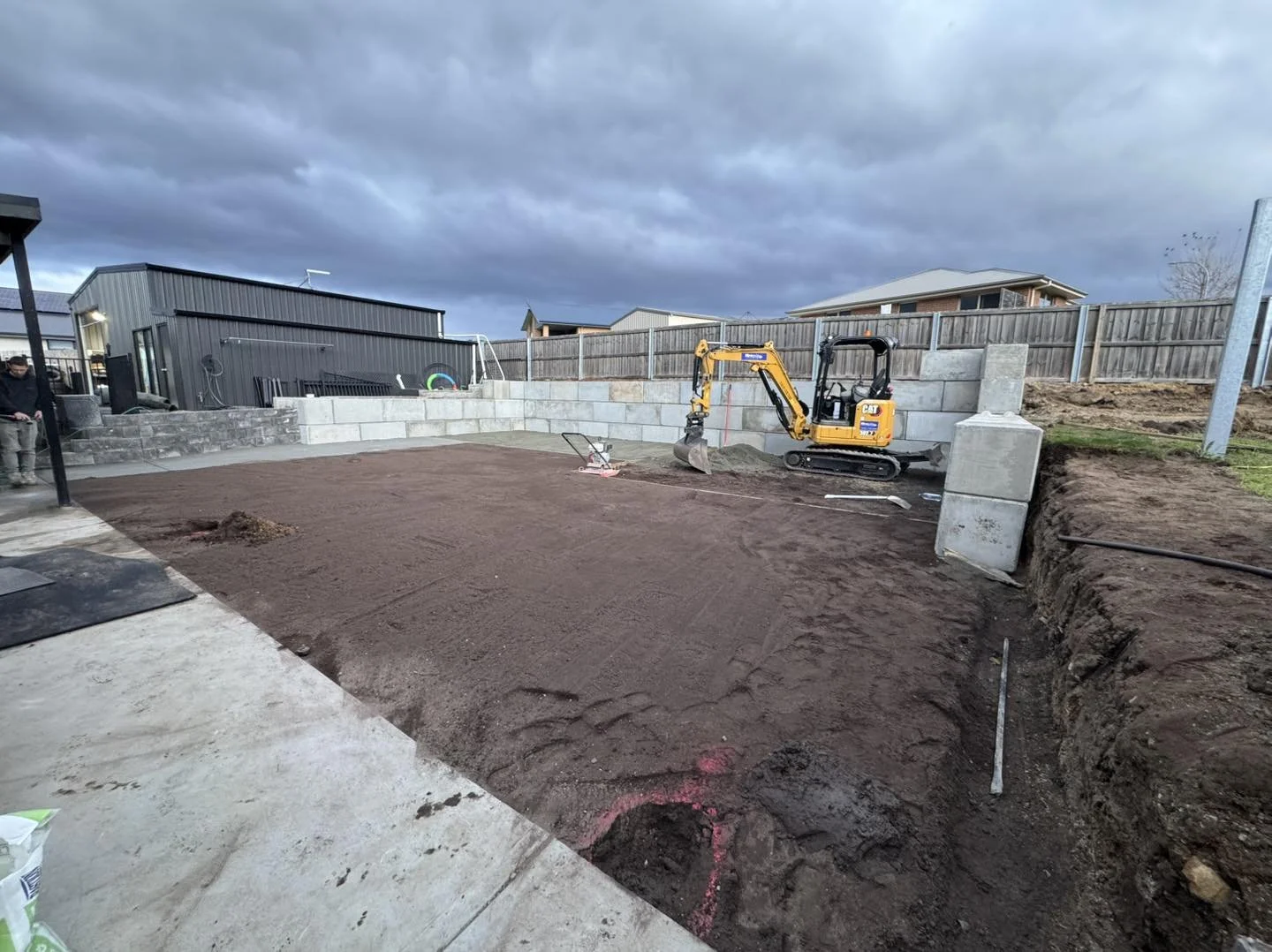Construction site with a mini excavator moving dirt, an area prepared with a dirt surface, and a concrete retaining wall in the background, with cloudy skies overhead.