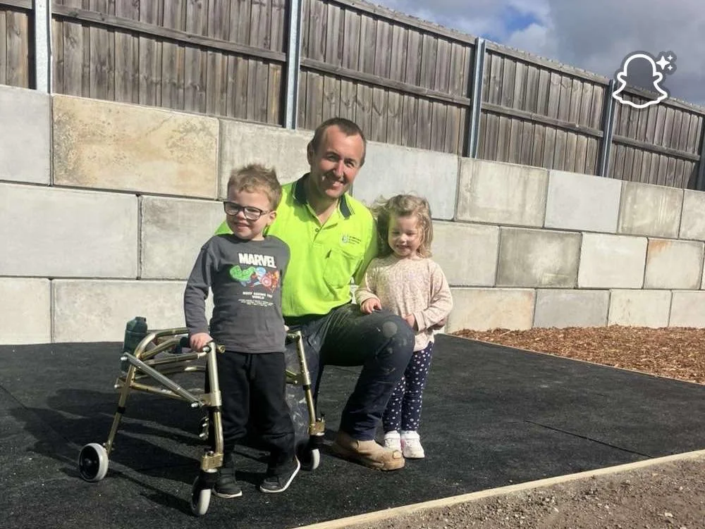 A man kneeling on a paved ground with two young children, one boy with a walker and one girl, outdoors with a wooden fence and concrete blocks behind them.