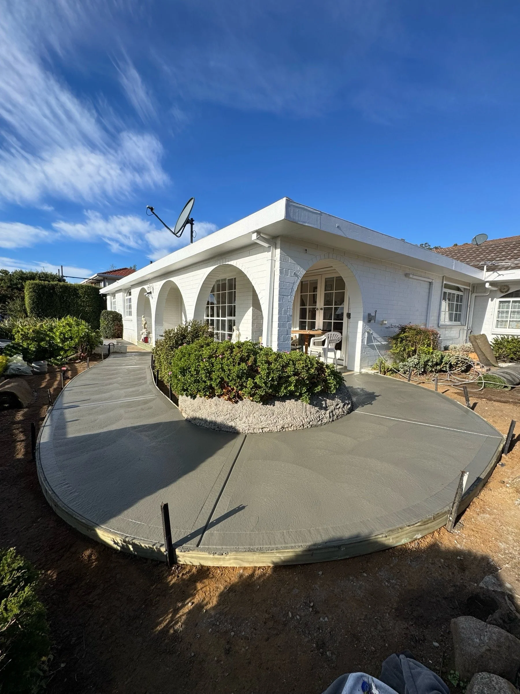 A white house with a covered porch, multiple windows, and two satellite dishes on the roof. In front of the house, there is a newly poured concrete sidewalk and a raised round garden bed with green bushes. The sky is partly cloudy with blue patches.