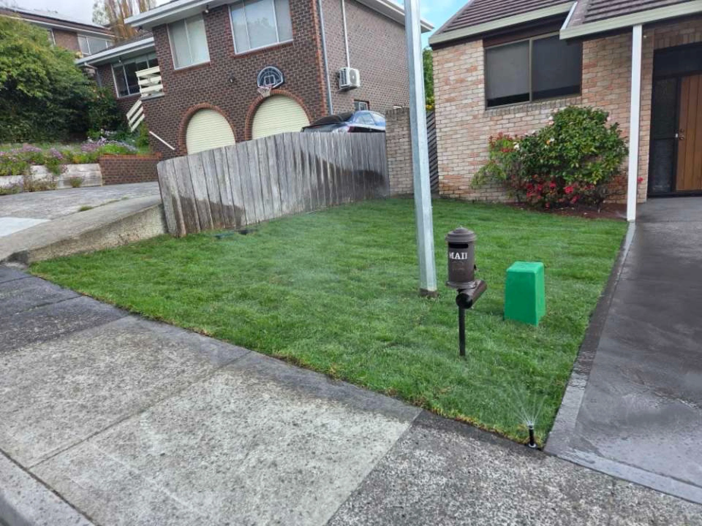 A small front yard with freshly cut green grass, bordered by a concrete sidewalk and driveway. A black outdoor sprinkler is watering the grass at the edge. A black mailbox with the name 'MAY' is mounted on a post near the sidewalk. There are two utility poles, one white and one yellow, in the yard. Behind the yard, there is a brick house on the right with a door, window, and bushes, and a neighboring brick structure on the left with a garage door, window, and a small basketball hoop. A weathered wooden fence separates the properties.