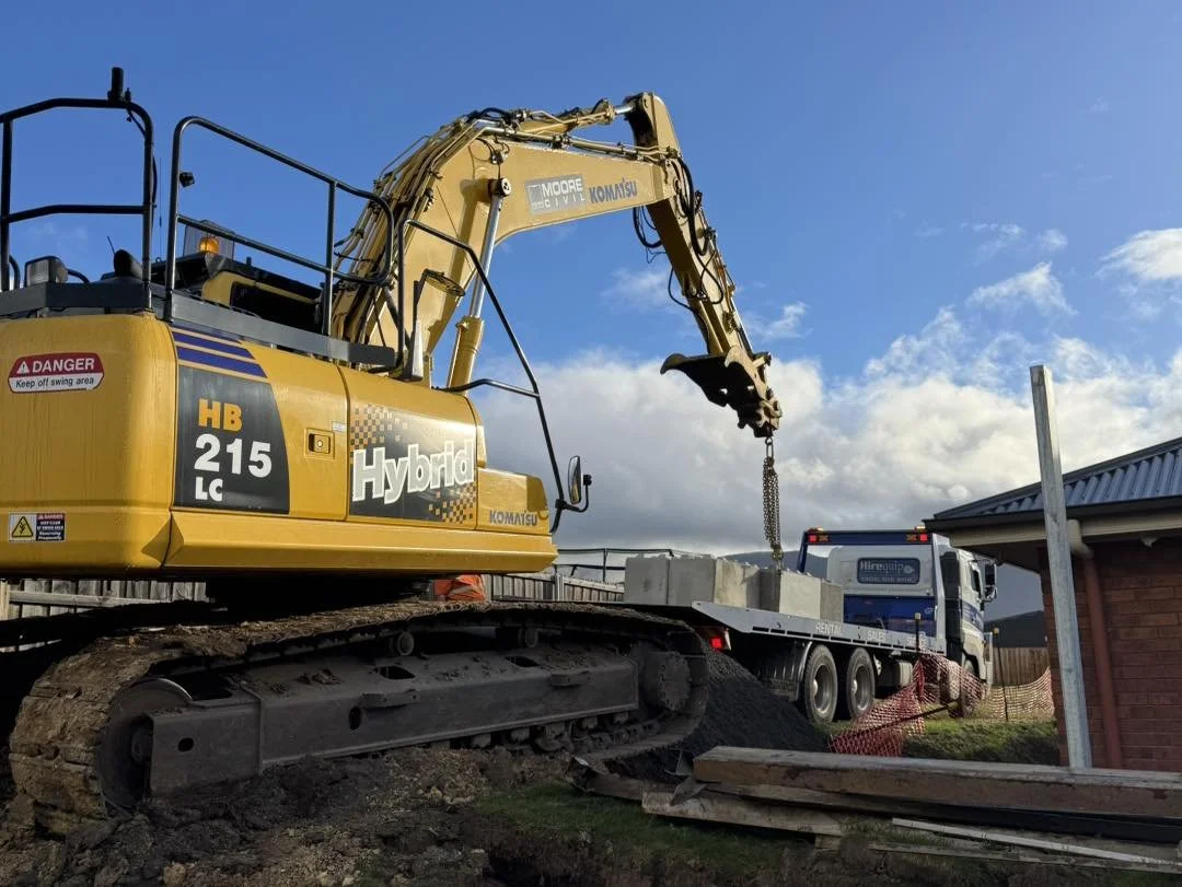 Construction site with a yellow hybrid excavator lifting concrete blocks onto a flatbed truck near a building with a brick wall. The sky is partly cloudy.