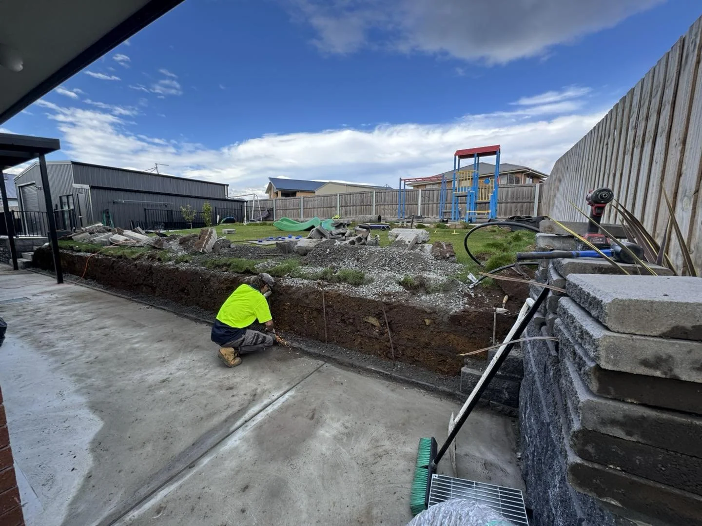 A construction worker in a yellow safety vest and gray pants crouches near a trench in a backyard, working on the foundation or plumbing. The backyard is under renovation, with piles of dirt, gravel, and construction tools visible. In the background,