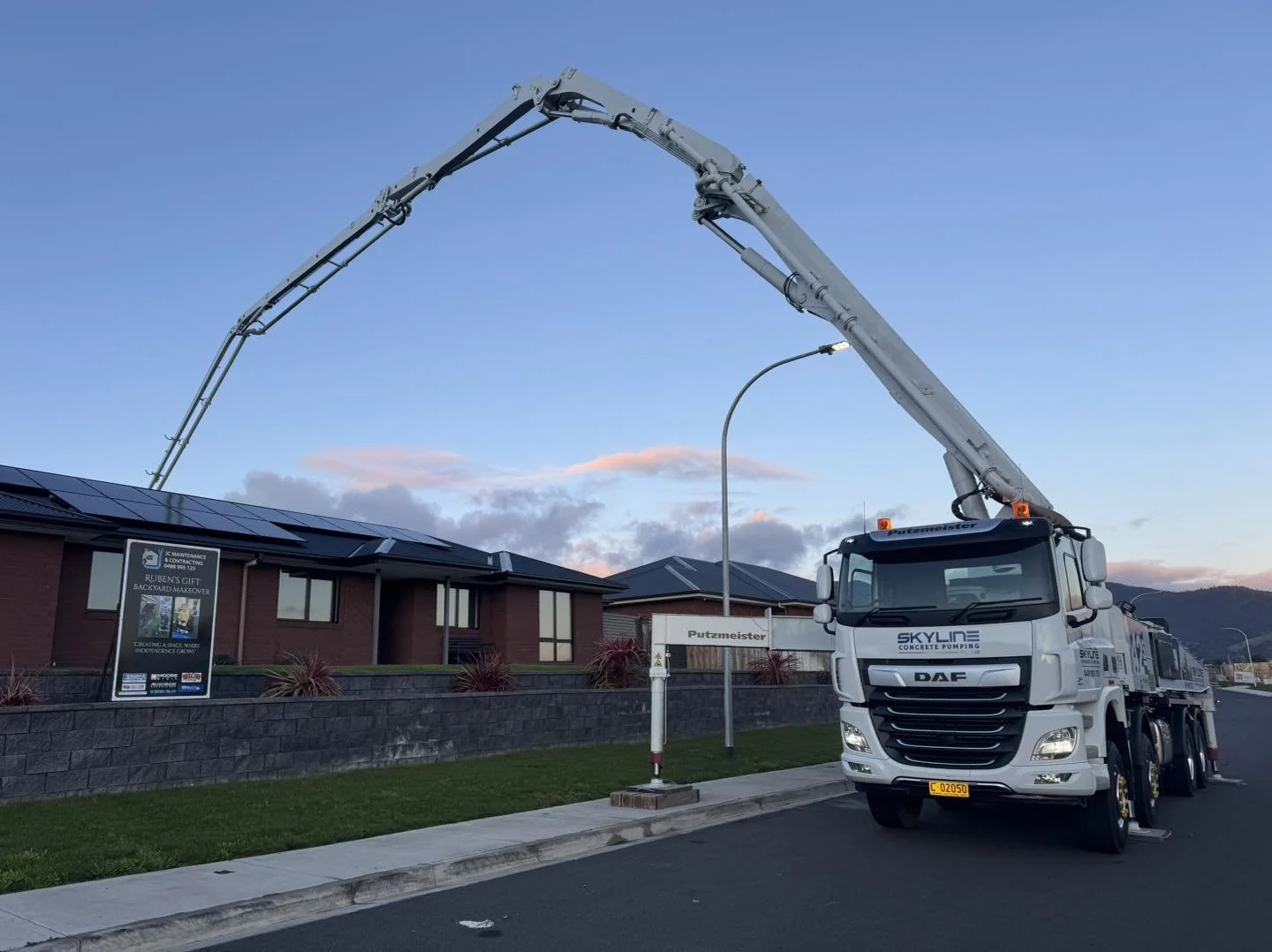 A large white truck with a long boom arm working on a building's roof. The truck is parked on the street, and a sign for Putzmeister Concrete Pumping is visible. The building has solar panels on the roof, and there is a sign that reads 'Ruben's Cift 