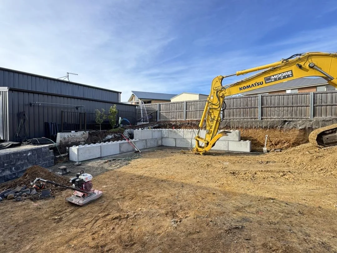 Construction site with a yellow excavator and concrete blocks, fenced backyard, and clear sky.