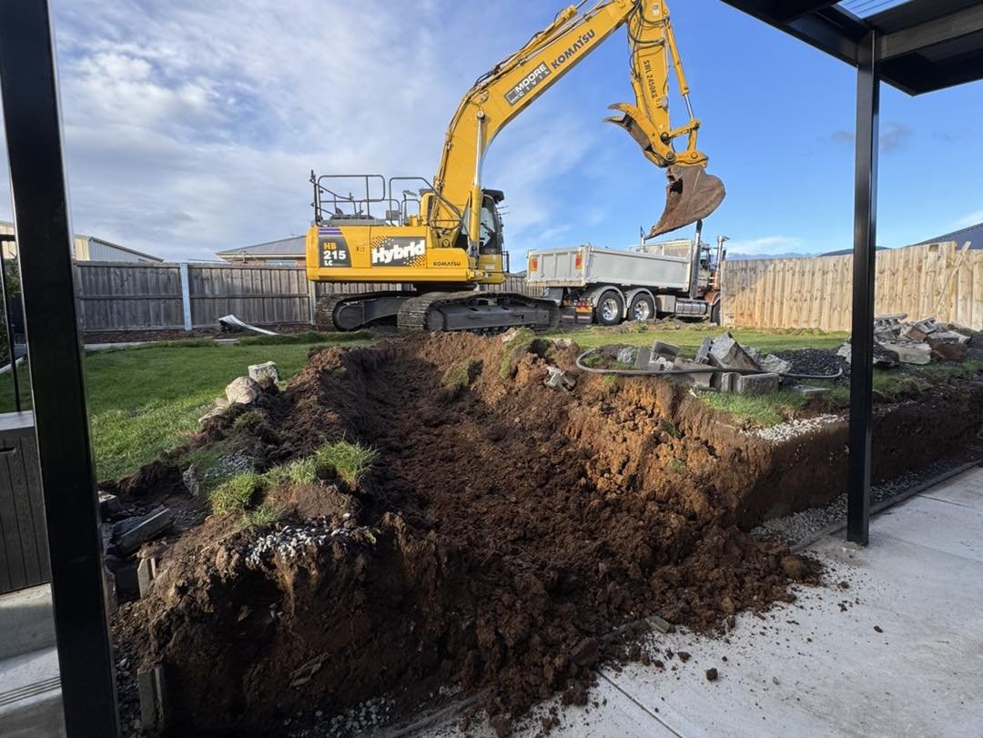 A yellow Komatsu excavator digging a trench in a backyard with grass and a wooden fence, with a dump truck nearby.