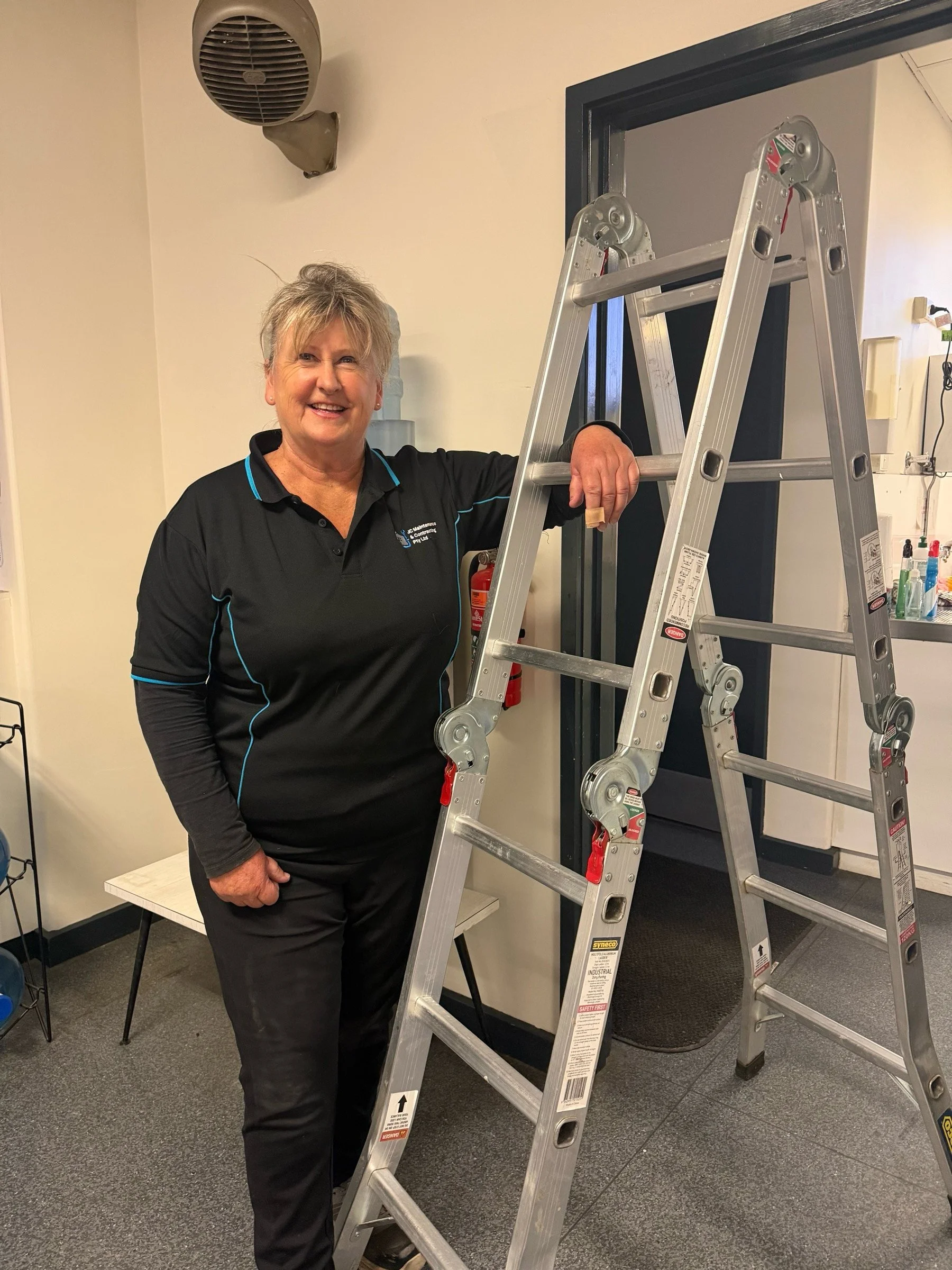 A smiling woman with short blonde hair standing next to a ladder indoors. She is wearing a black polo shirt with blue accents.