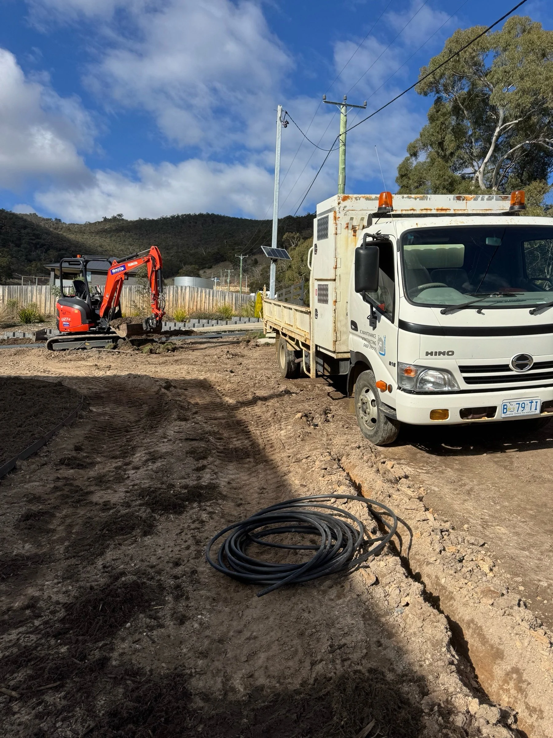 Construction site with a small orange excavator, a white utility truck, and coiled black cable on dirt ground under a partly cloudy sky.