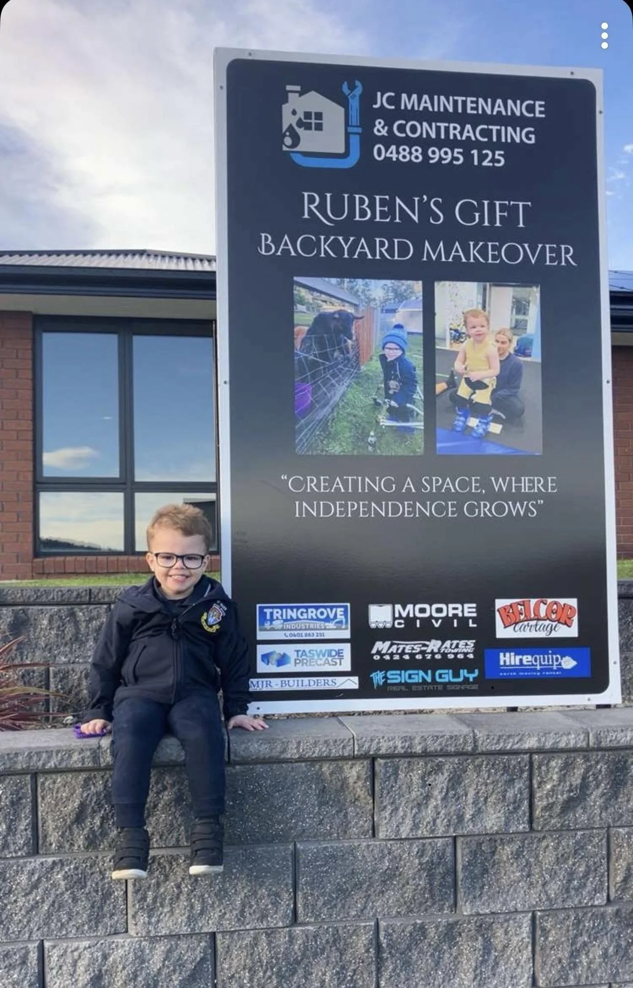 Child sitting on a stone wall next to a large sign advertising Ruben's Backyard Makeover. The sign includes images of children and various company logos.