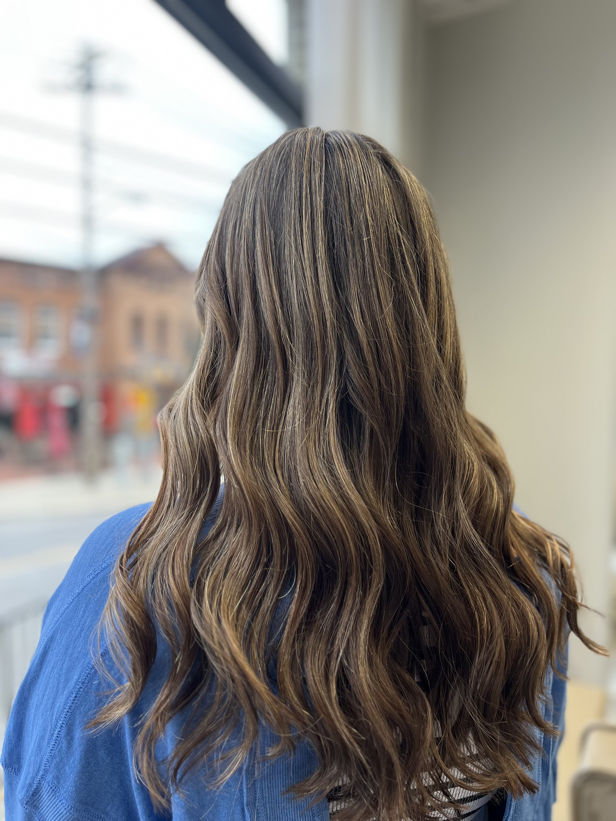 Close-up of a woman with long, wavy, light brown hair wearing a blue shirt, looking out a window with blurred buildings outside.