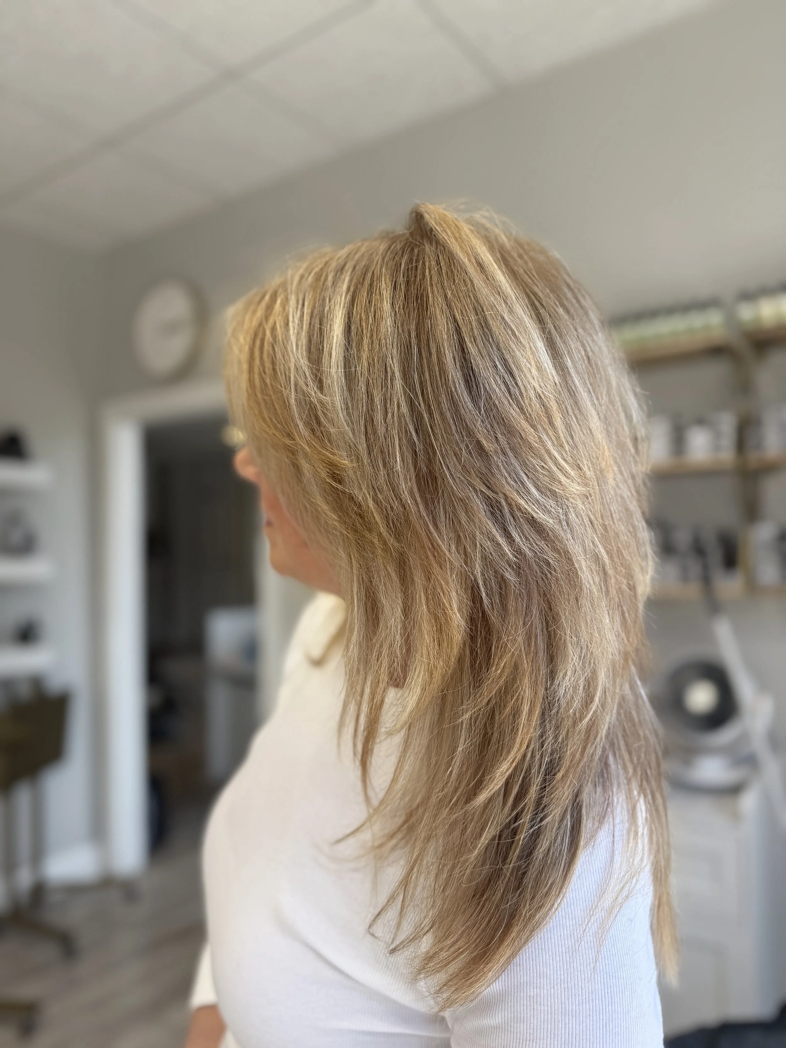 Close-up of a woman with honey-blonde hair, wearing a white top, in a room with shelving and a clock in the background.