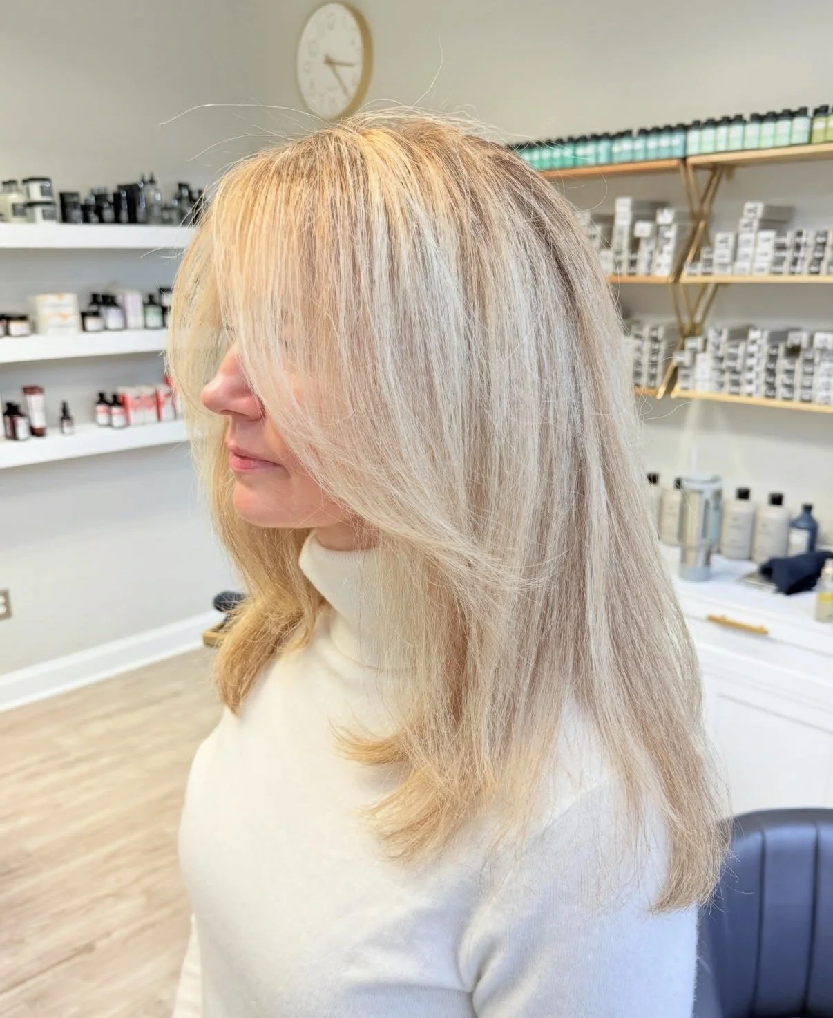 A woman with long, blonde hair in a salon, facing sideways, with shelves of hair products in the background.