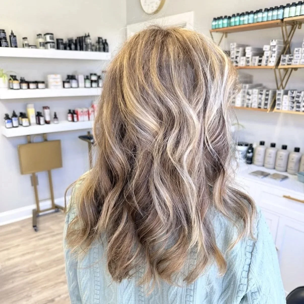 Back of a woman with blonde, wavy hair in a salon or spa setting, with shelves of hair and skincare products in the background.