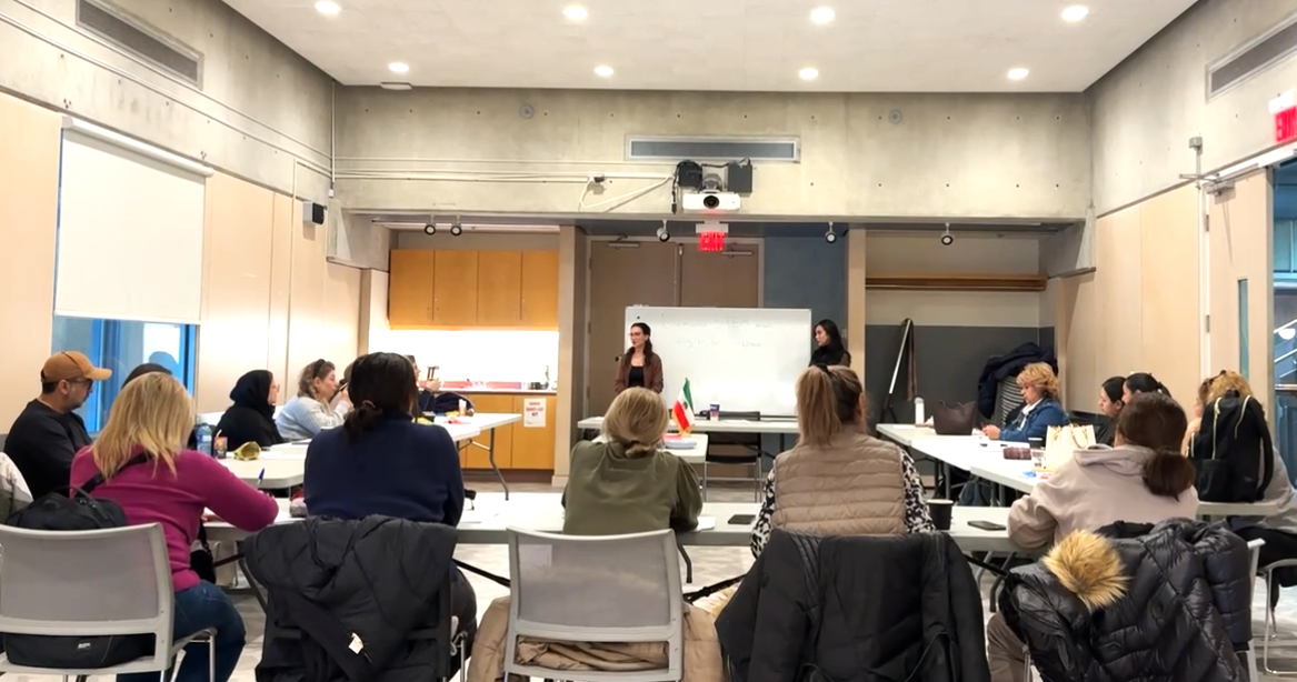 A group of people attending a presentation in a conference room with a whiteboard at the front. Two women are speaking at the front of the room, one of them is standing near a flag.