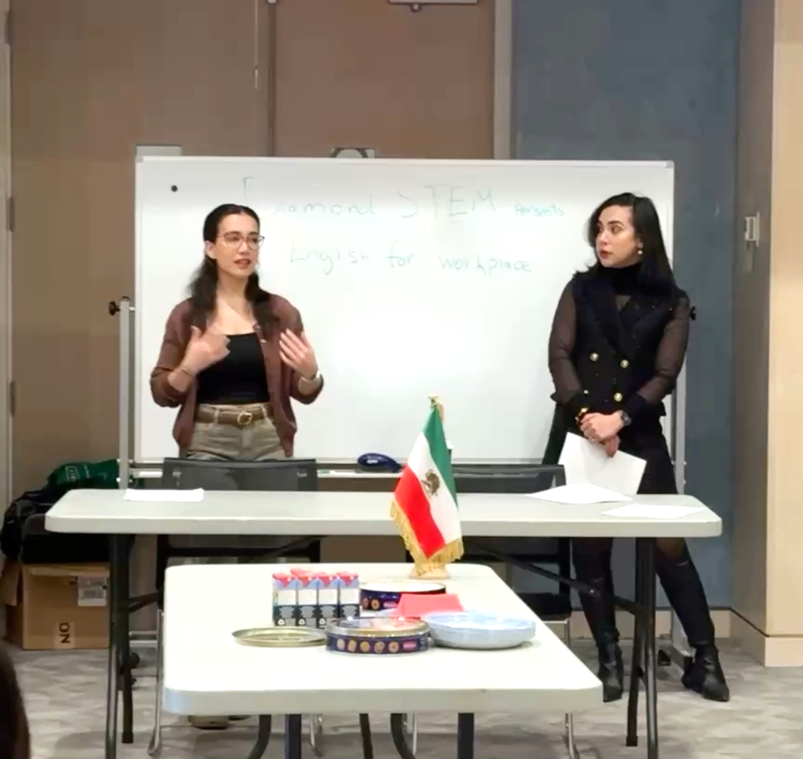 Two women standing in front of a whiteboard during a workshop, with an American flag on the table in front of them. The whiteboard has writing that says 'Diamond STEM presents English for Workshop.'