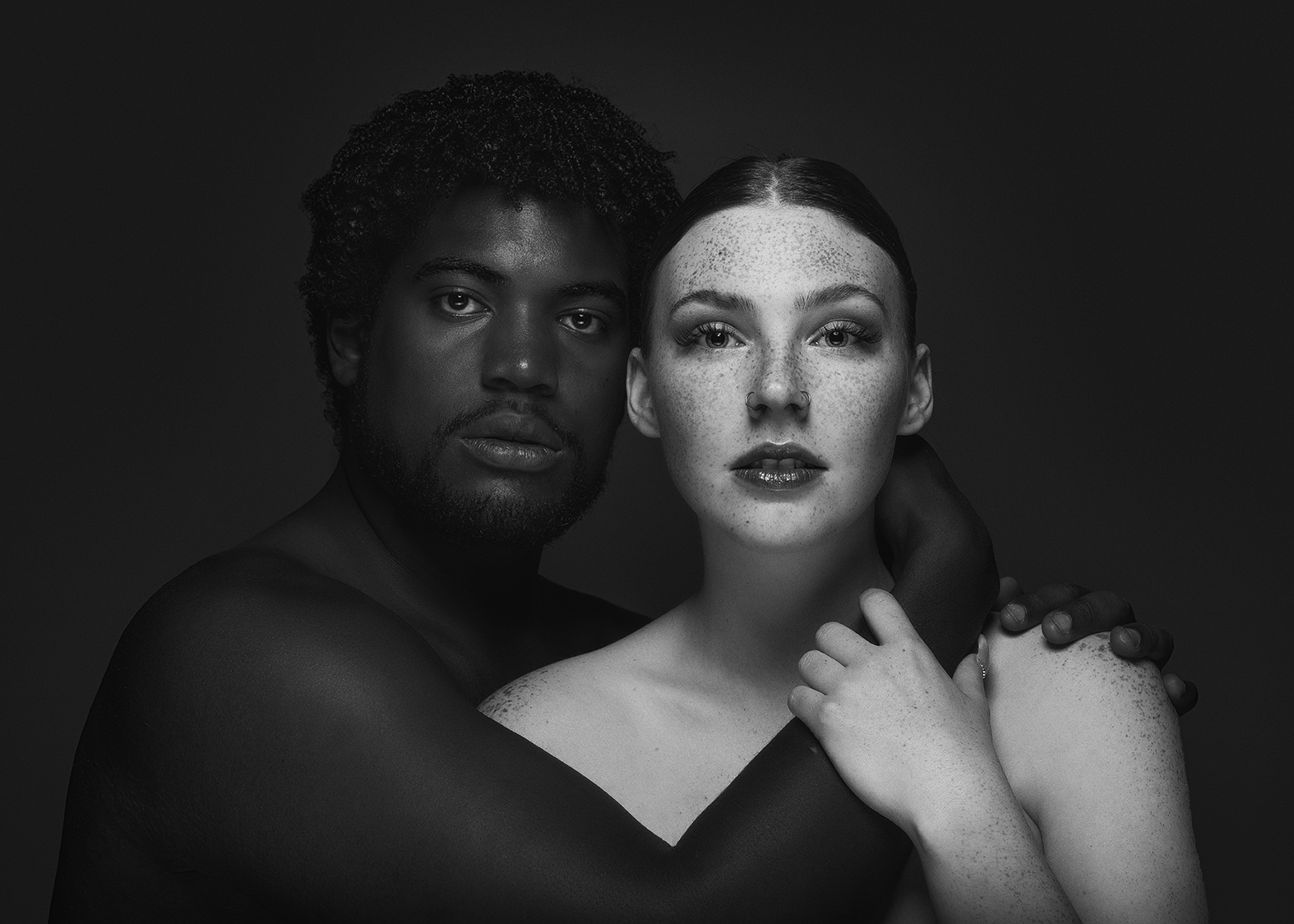 Black and white portrait of a Black man and a white woman with freckles, both looking at the camera. The man has his arm around the woman's shoulder, and she has her hand on his arm, with a neutral background.