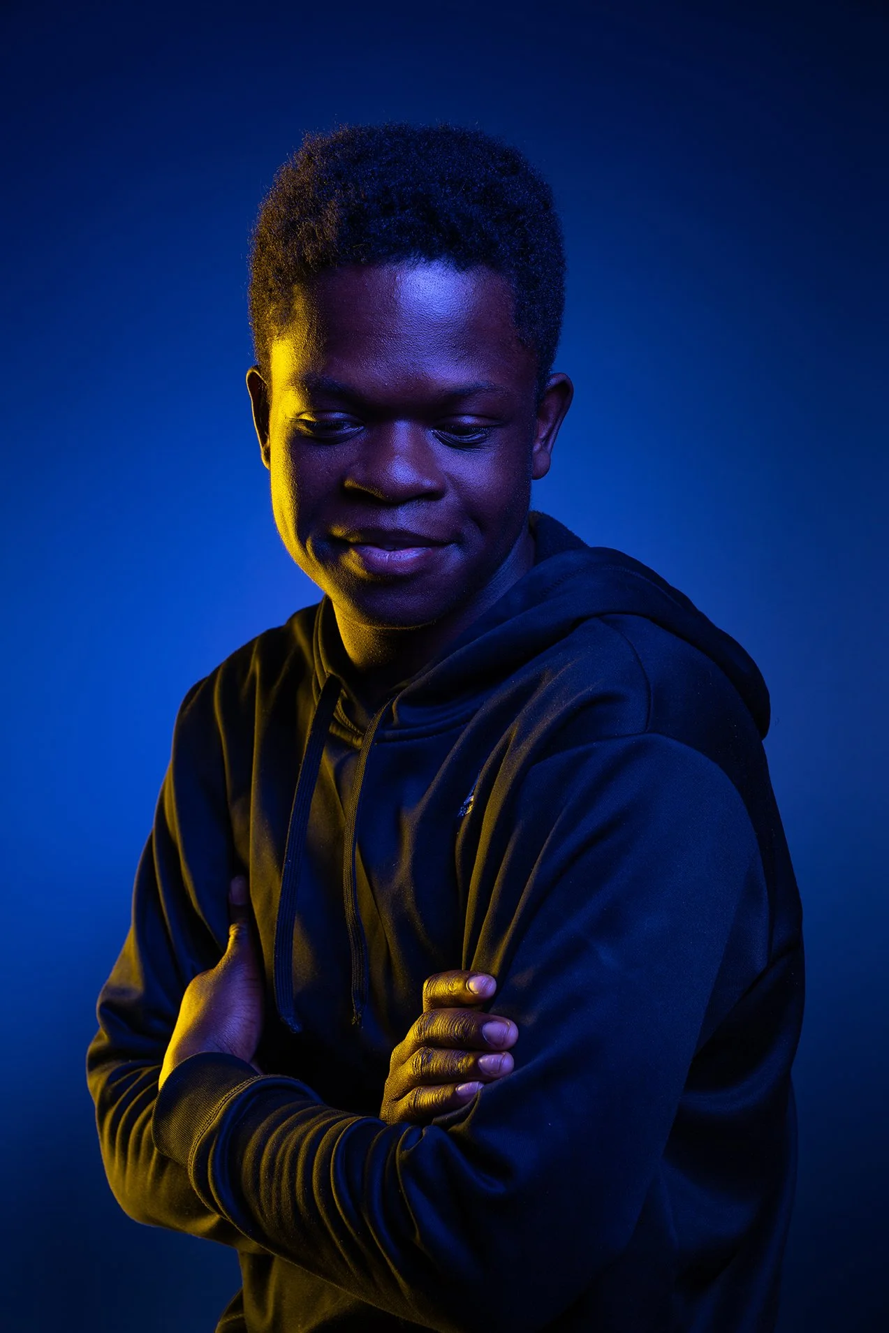 A young man standing with arms crossed against a dark blue background, illuminated with bluish and yellow lighting.
