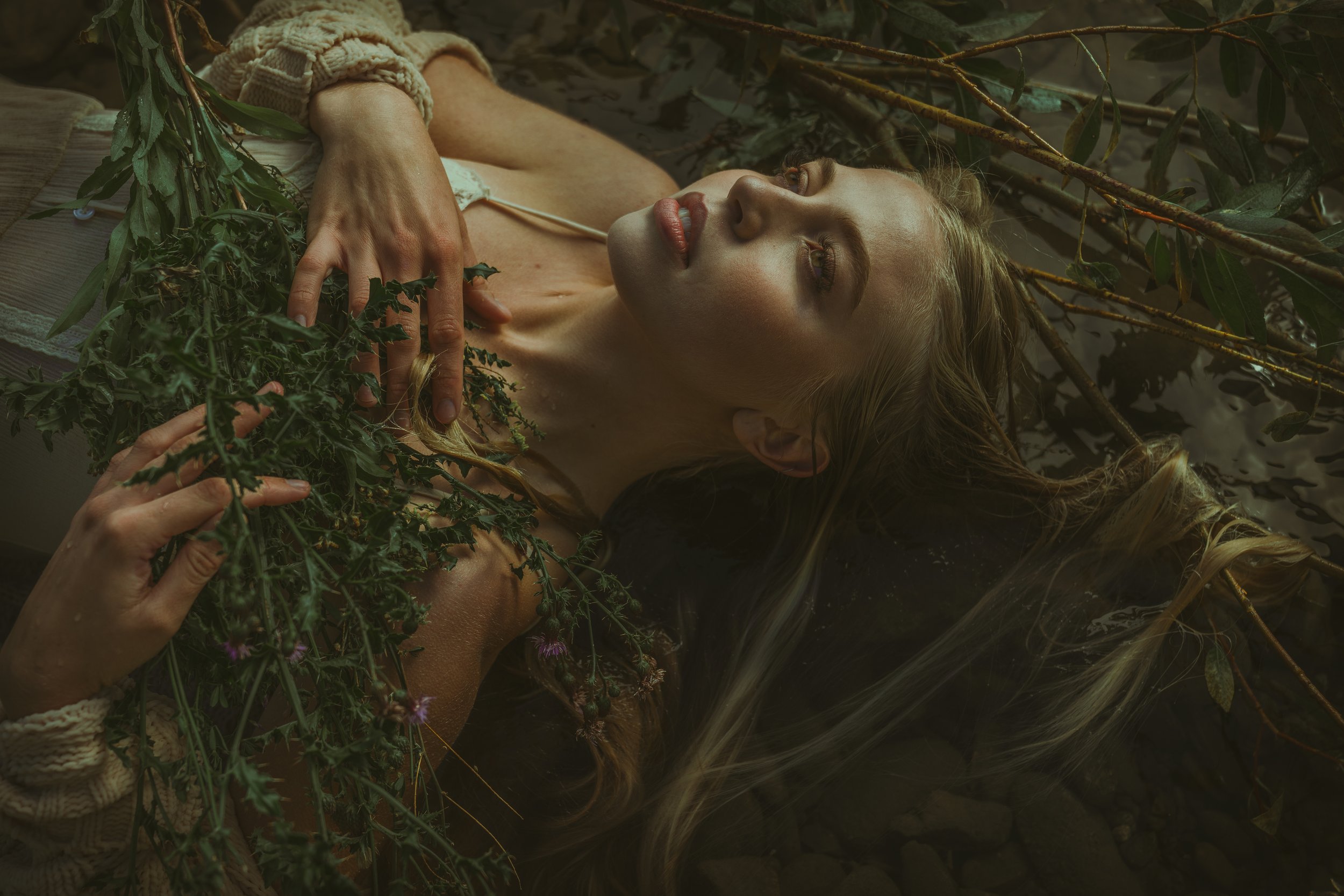 A woman with long blonde hair reclining in water surrounded by green leaves and branches, with a contemplative expression.
