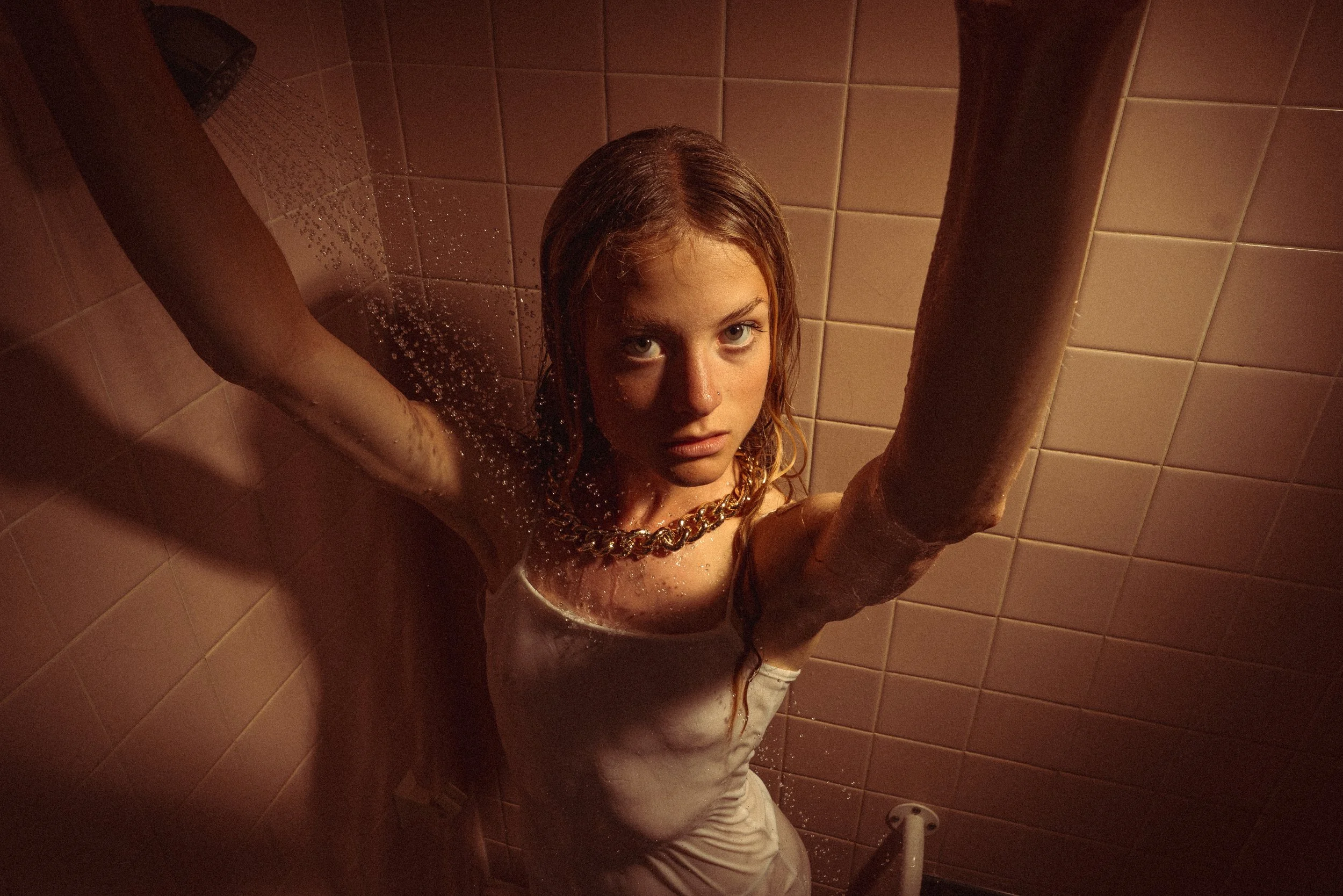 A woman with wet hair in a shower, looking up at the camera with a serious expression, surrounded by pink tiles.