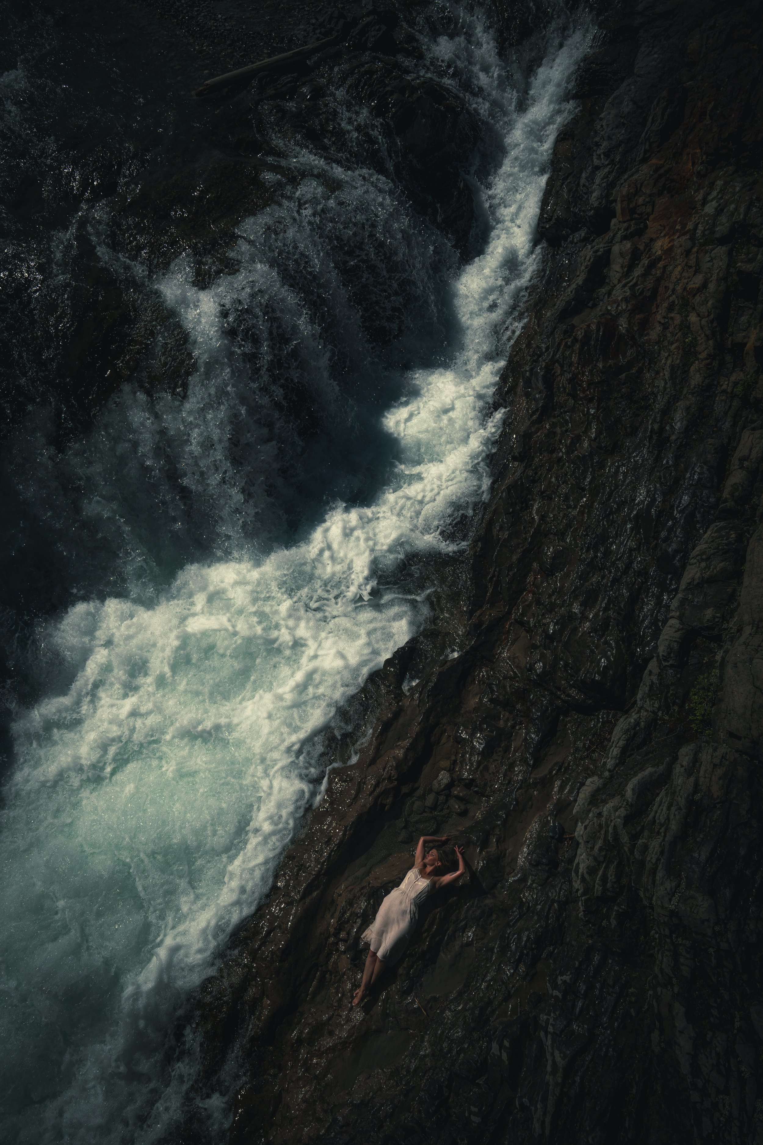 A woman lying on a rock next to a fast-flowing river waterfall with dark, jagged rocks around her.