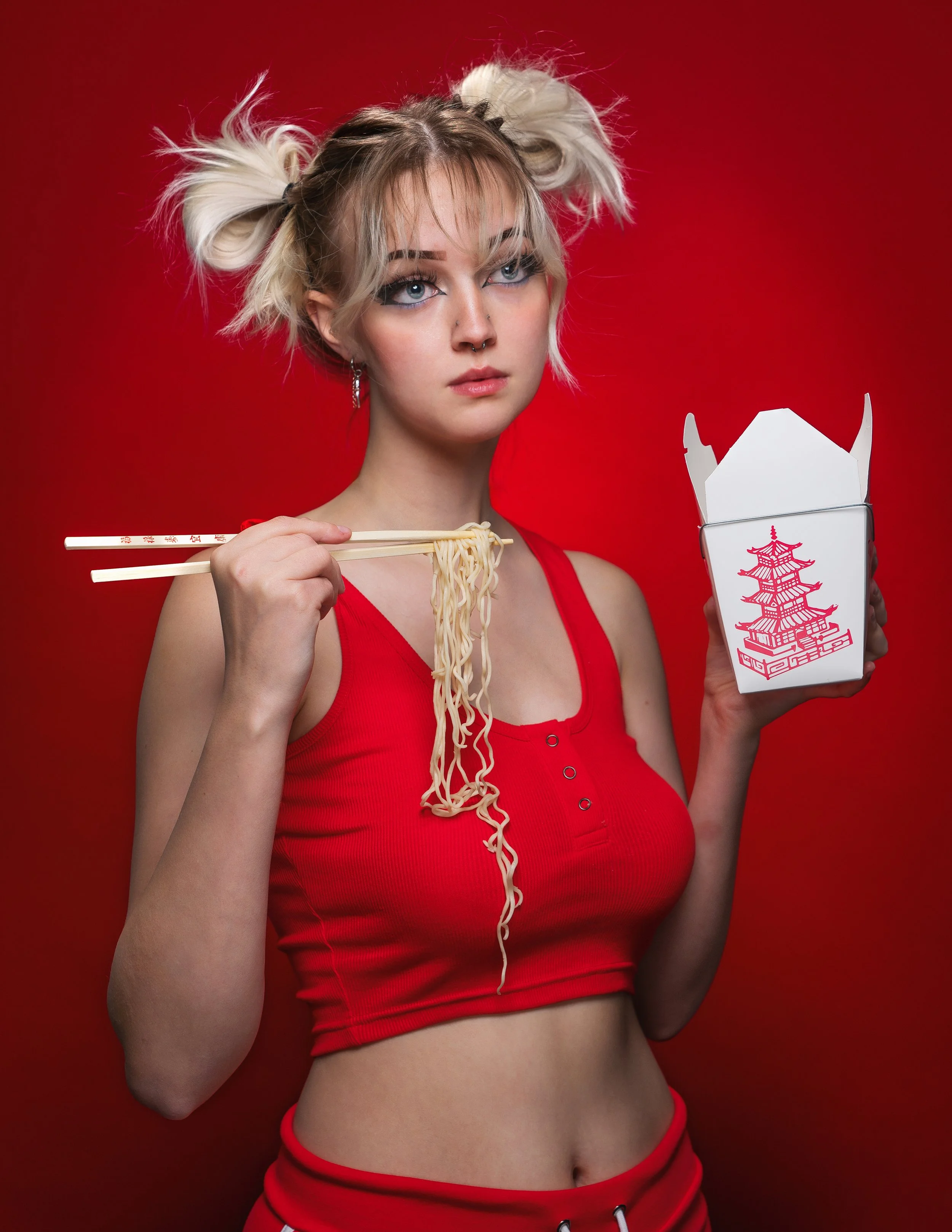 Young woman with blonde hair in pigtails, wearing a red tank top, holding chopsticks with noodles in one hand and a takeout box with a pagoda illustration in the other, against a red background.