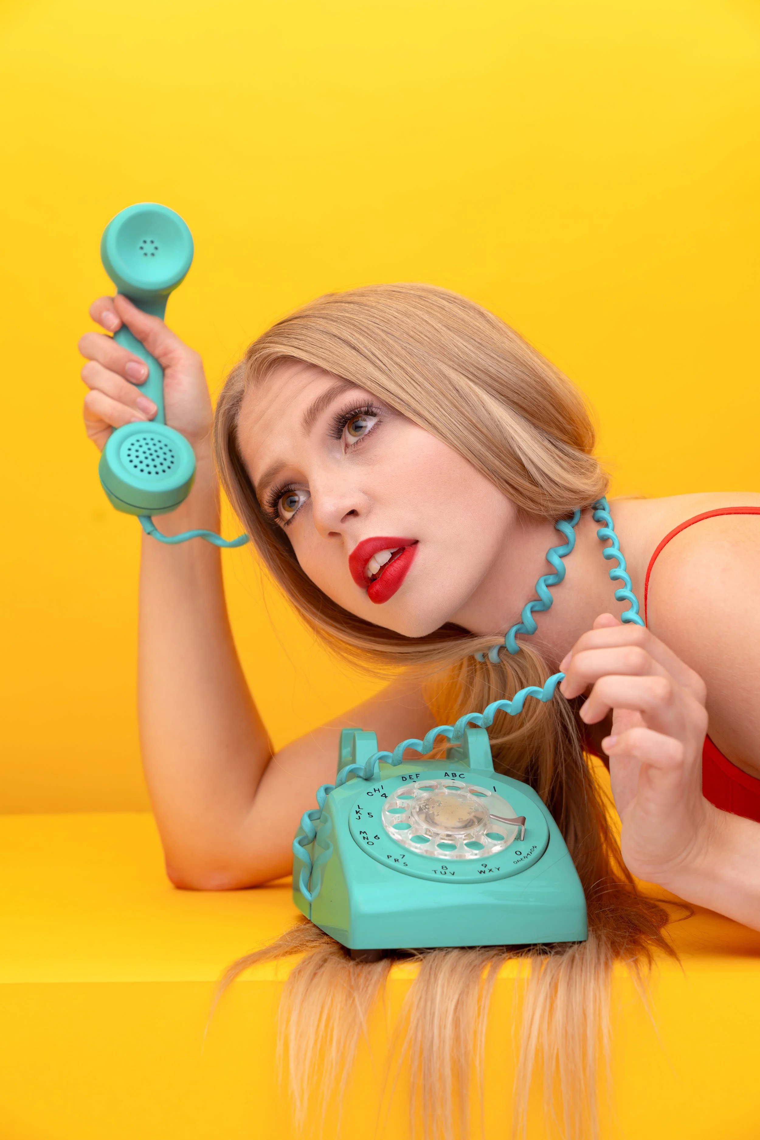 A woman with red lipstick and blonde hair, wearing a red top, is lying on a yellow surface. She is holding a turquoise vintage rotary phone with the receiver in her left hand, resting her head on her right arm, and looking up.