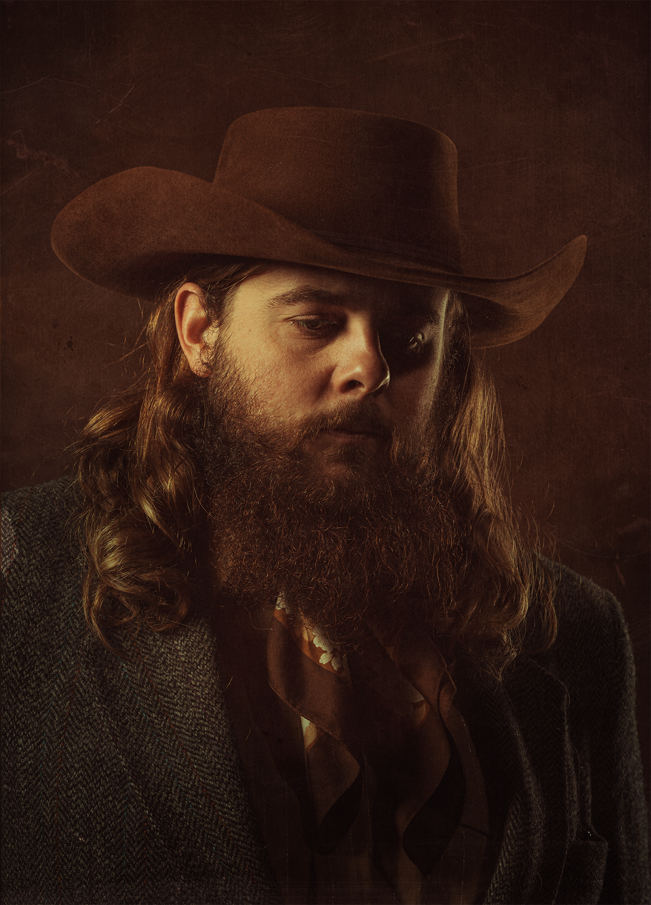 A man with long hair and beard wearing a brown cowboy hat, plaid shirt, and dark jacket, looking downward against a dark background.