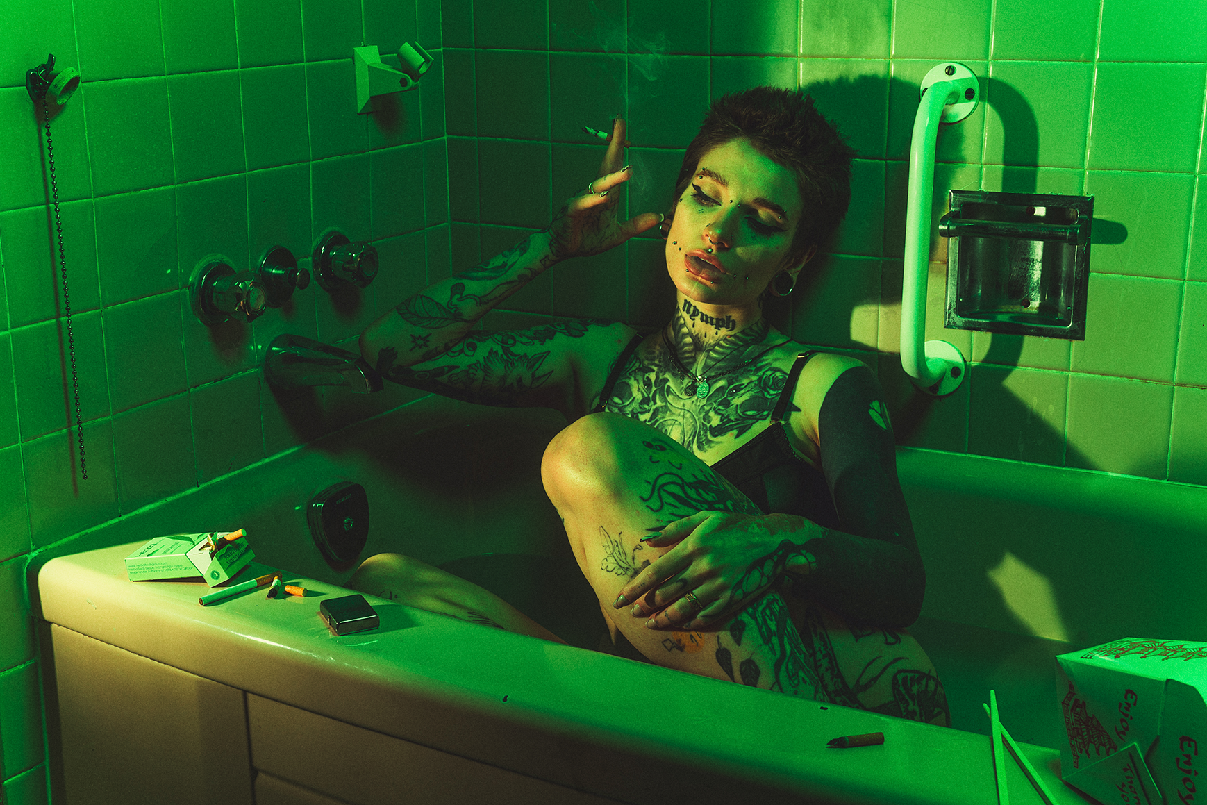 A tattooed woman with short hair in a bathtub with green lighting, smoking a cigarette, surrounded by cigarette packs and lighters.