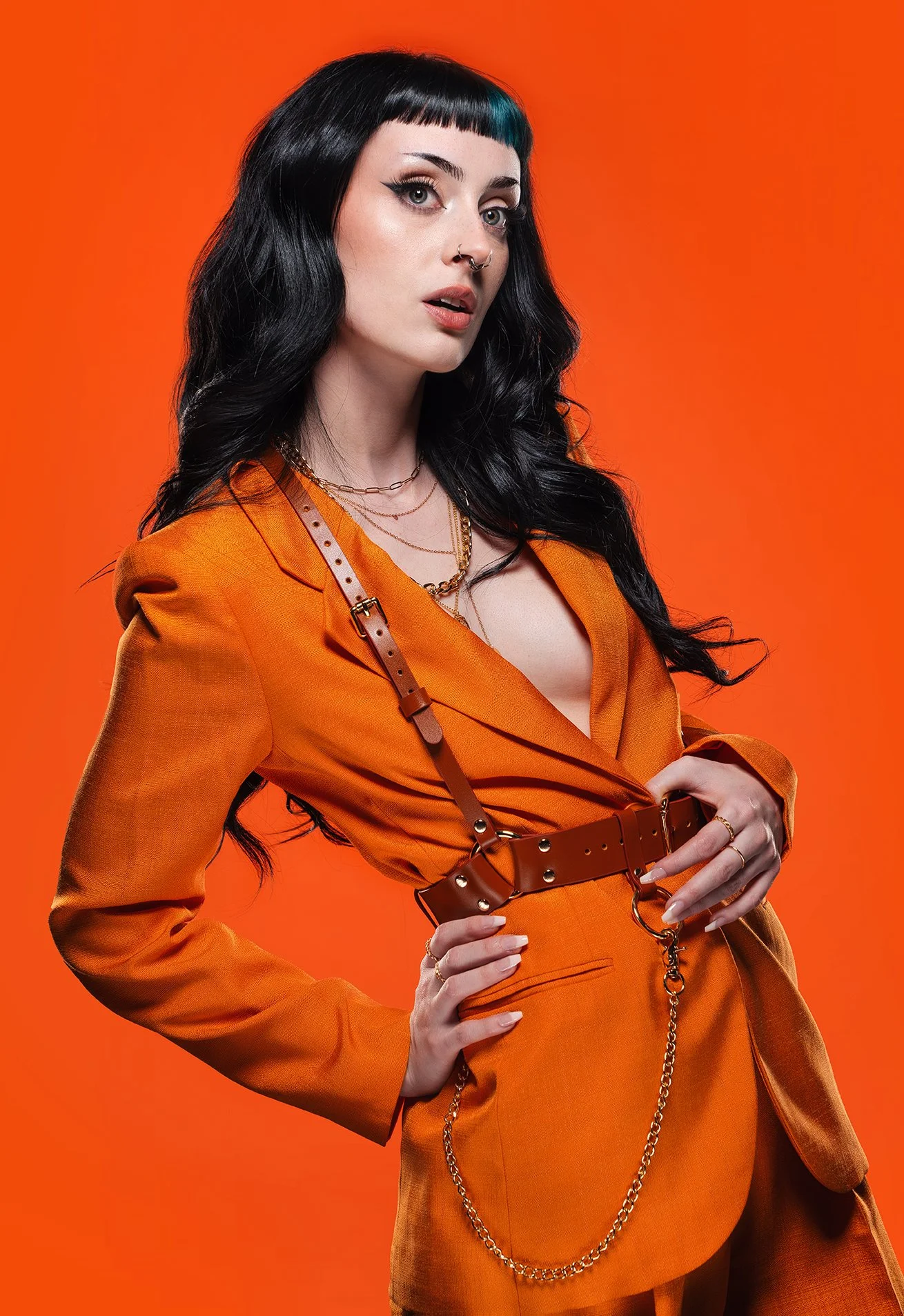 A woman with black hair, blue streaks, and fair skin, wearing an orange blazer with a matching belt and chain accessories, stands against an orange background.