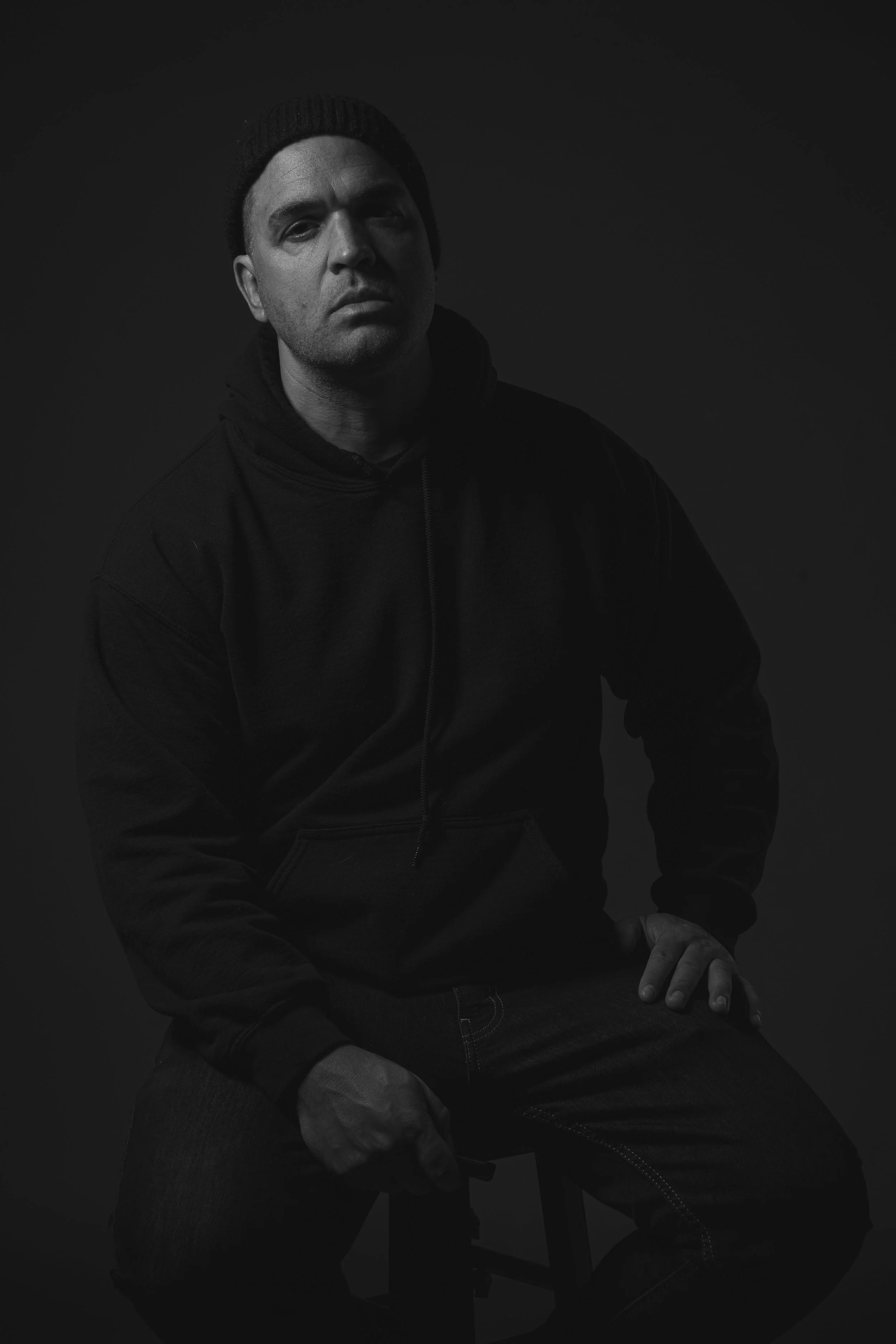 Black and white portrait of a man wearing a beanie, hoodie, and jeans, sitting on a stool against a dark background.