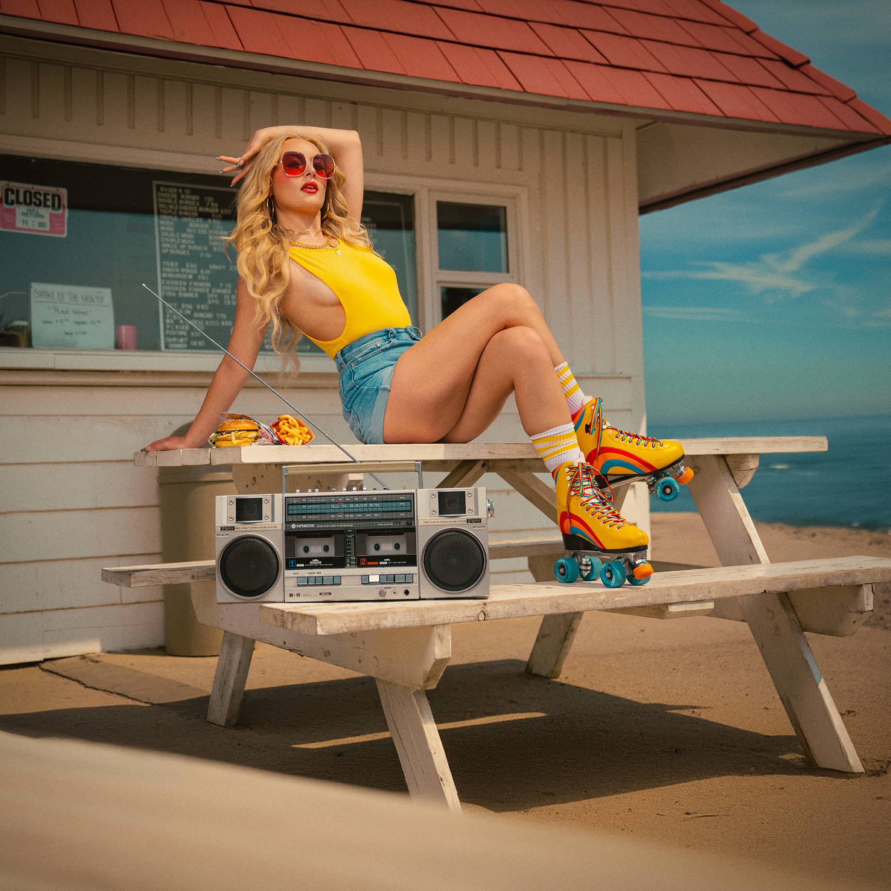 A woman sitting on a picnic table at the beach, wearing yellow tank top, denim shorts, yellow roller skates, and striped socks, with a boombox, holding a pair of sunglasses, with a food tray nearby, and a beachside building in the background.
