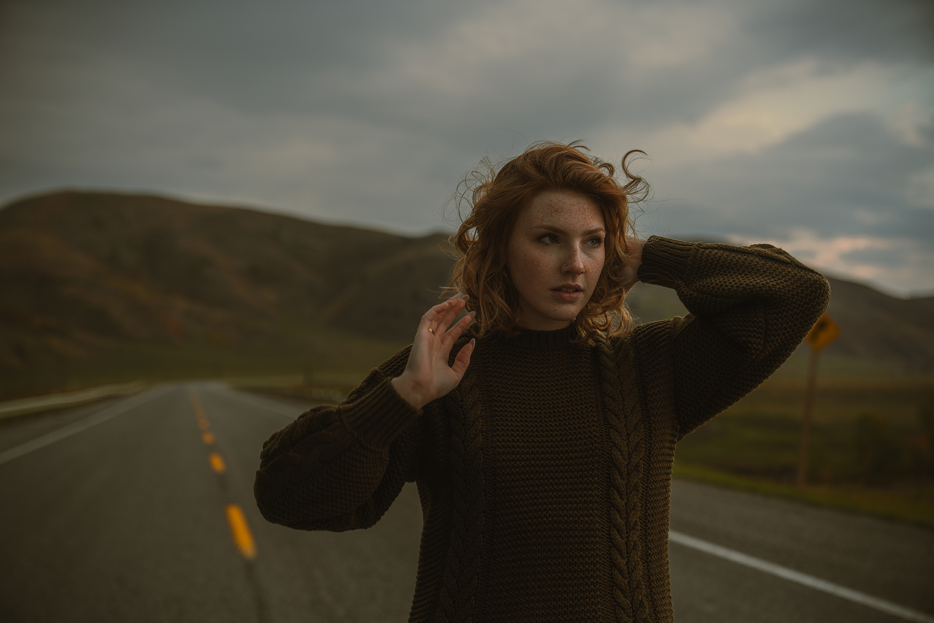 Young woman with red curly hair standing on an empty road with rolling hills in the background, wearing a brown knit sweater, under a cloudy sky.
