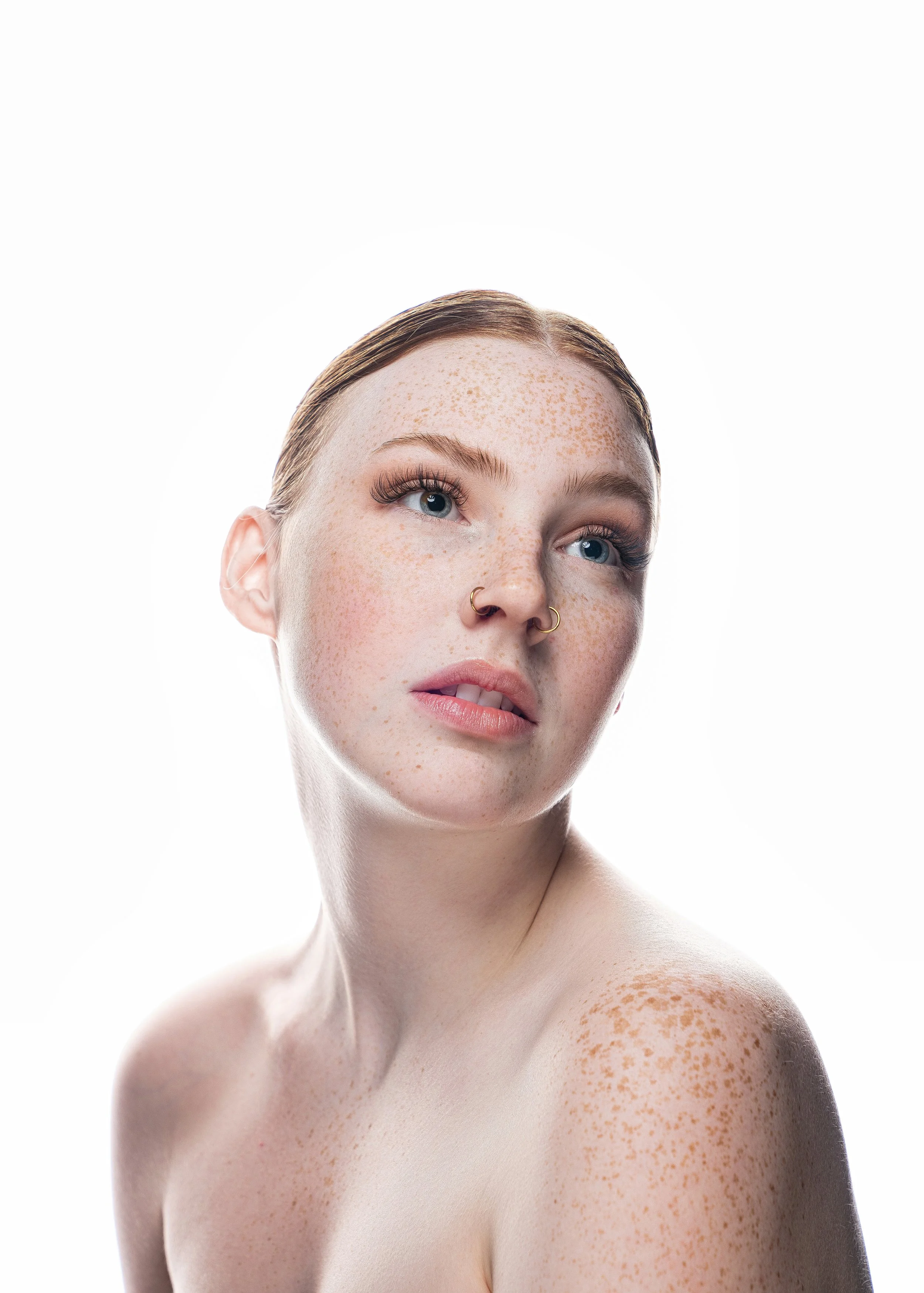 Close-up portrait of a young woman with red hair and freckles, looking slightly to the side, with natural makeup and a septum piercing, against a white background.